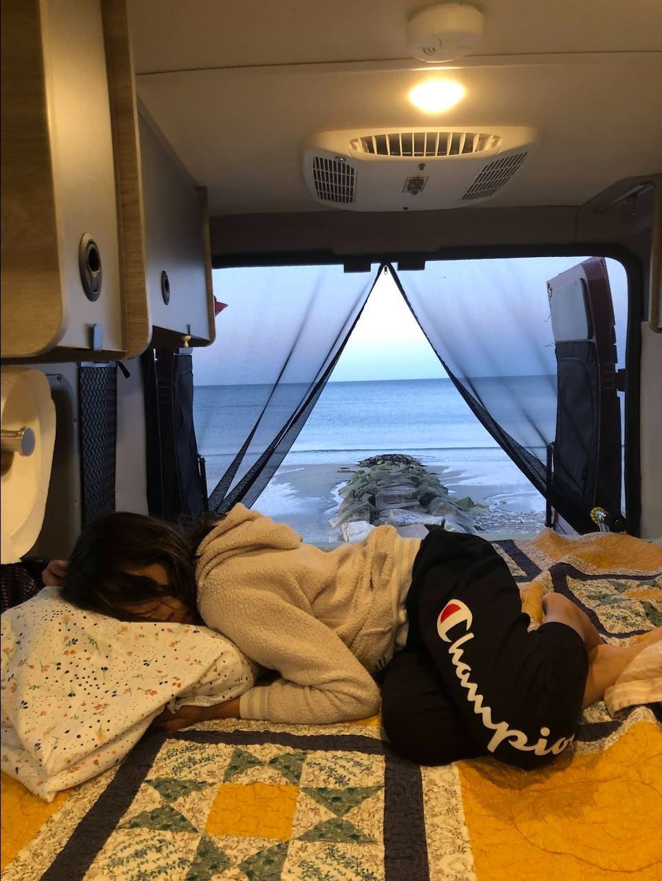 Inside an RV rental: A person sleeps on a cozy camper bed, facing a serene ocean and beach view. Ideal for nature travel.. Winnebago Solis 2021