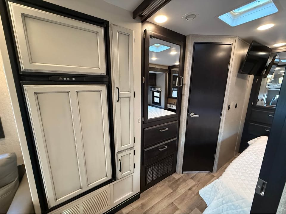 This RV rental's interior boasts a modern light-colored fridge, dark cabinetry, and a cozy bedroom with white bedding.. Entegra Coach Odyssey 2022