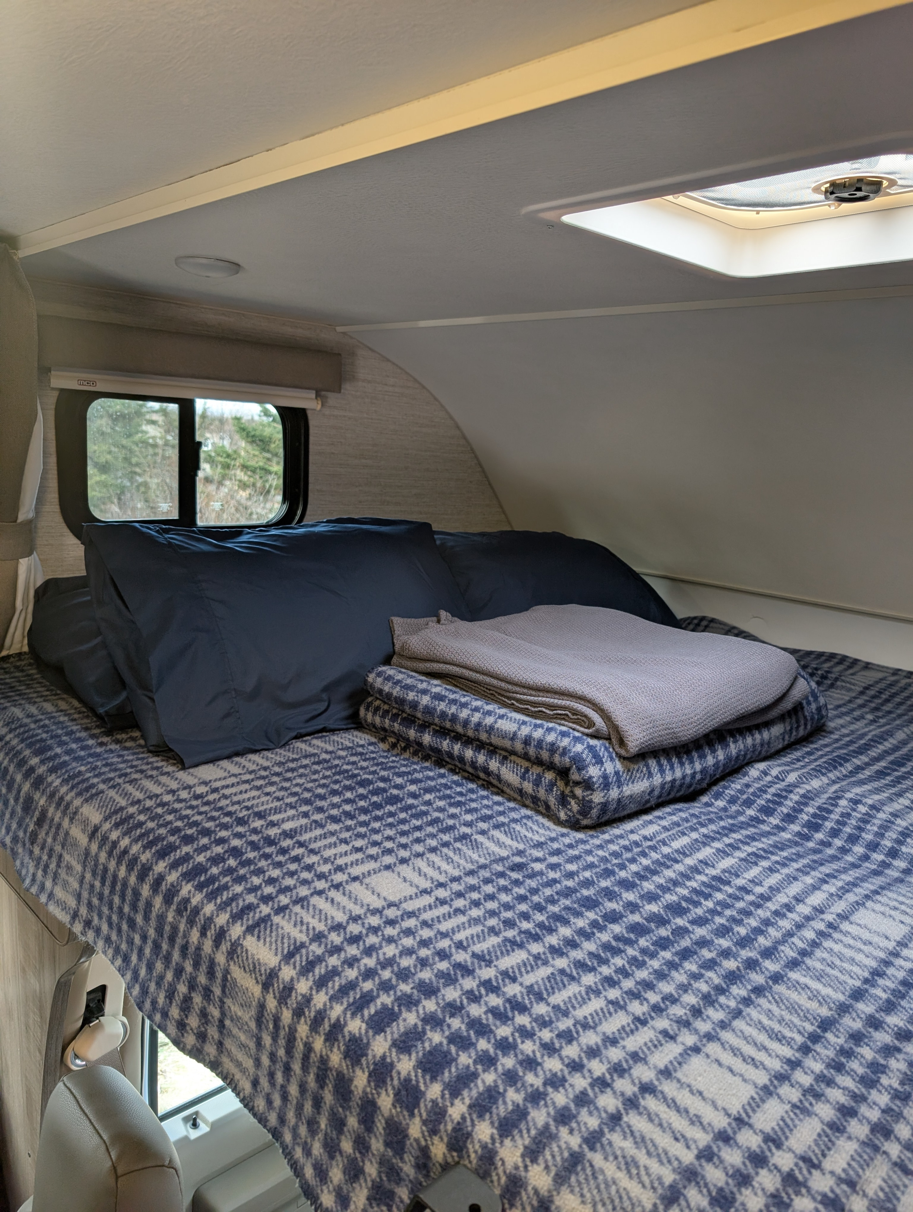 An RV rental's cozy bunk features a bed with blue plaid blankets, pillows, a window, and skylight.. Winnebago Minnie Winnie 2023