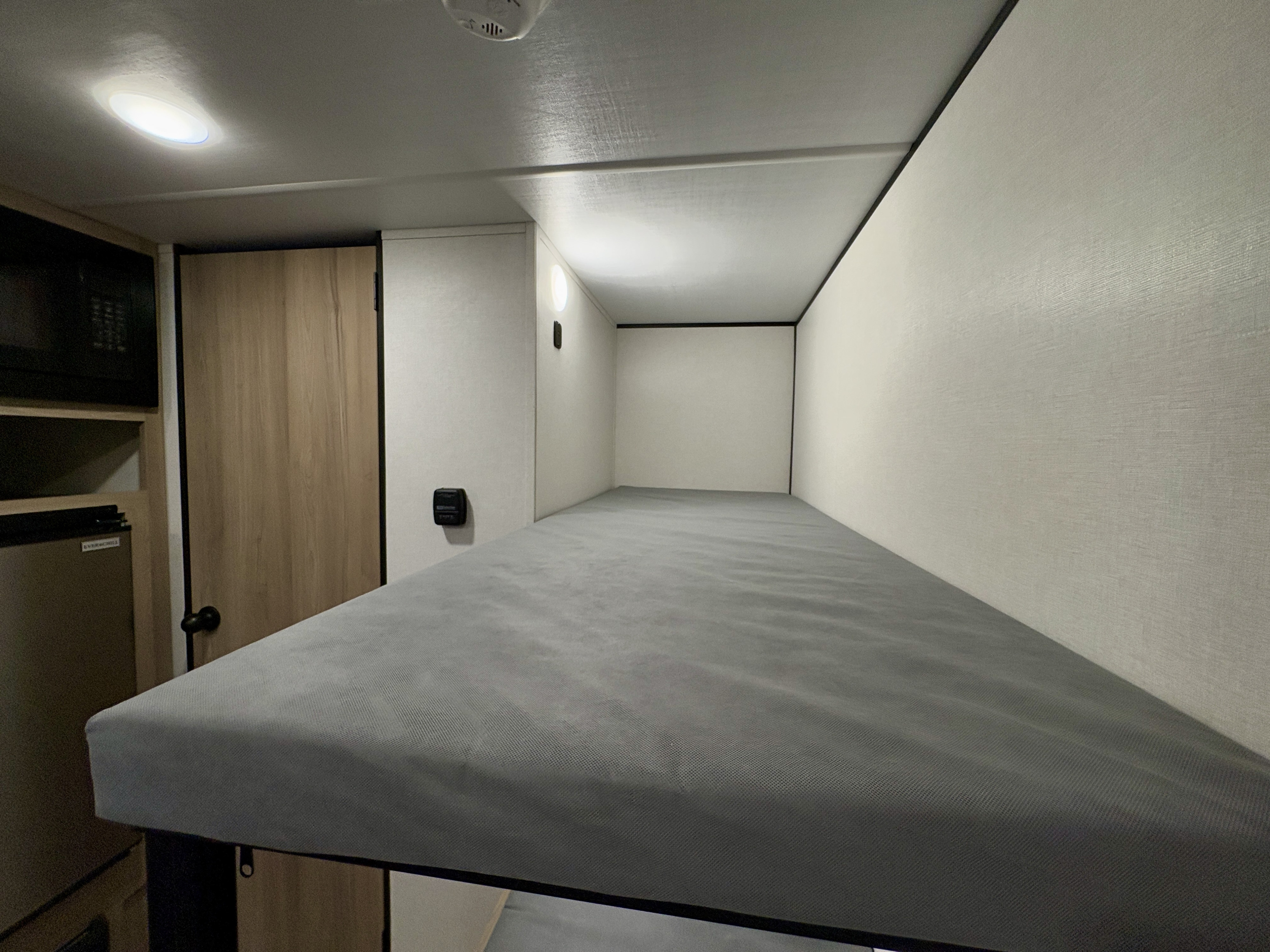 RV rental interior view from a grey-covered bunk bed, featuring a microwave, mini-fridge, and wooden door.. Coleman Keystone Light 17BX 2025