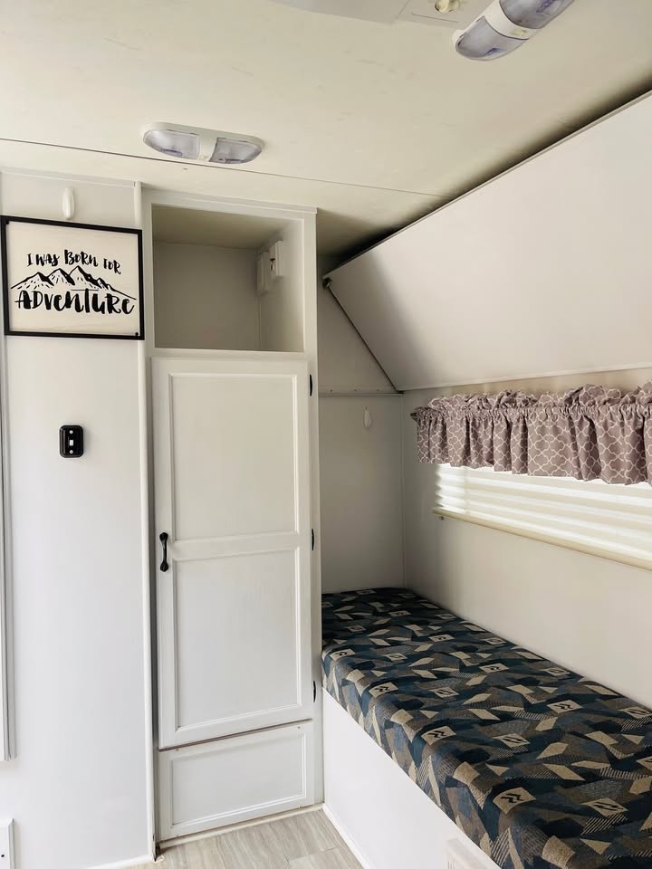 Cozy RV rental bunk area: white interior, patterned mattress, built-in storage, and adventure sign.. Cikira RV Classic Cruiser 2008