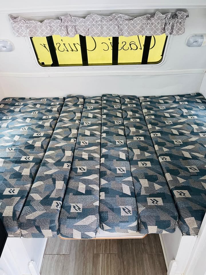 RV rental interior featuring a multi-segment bed with blue/grey geometric patterned cushions below a window.. Cikira RV Classic Cruiser 2008