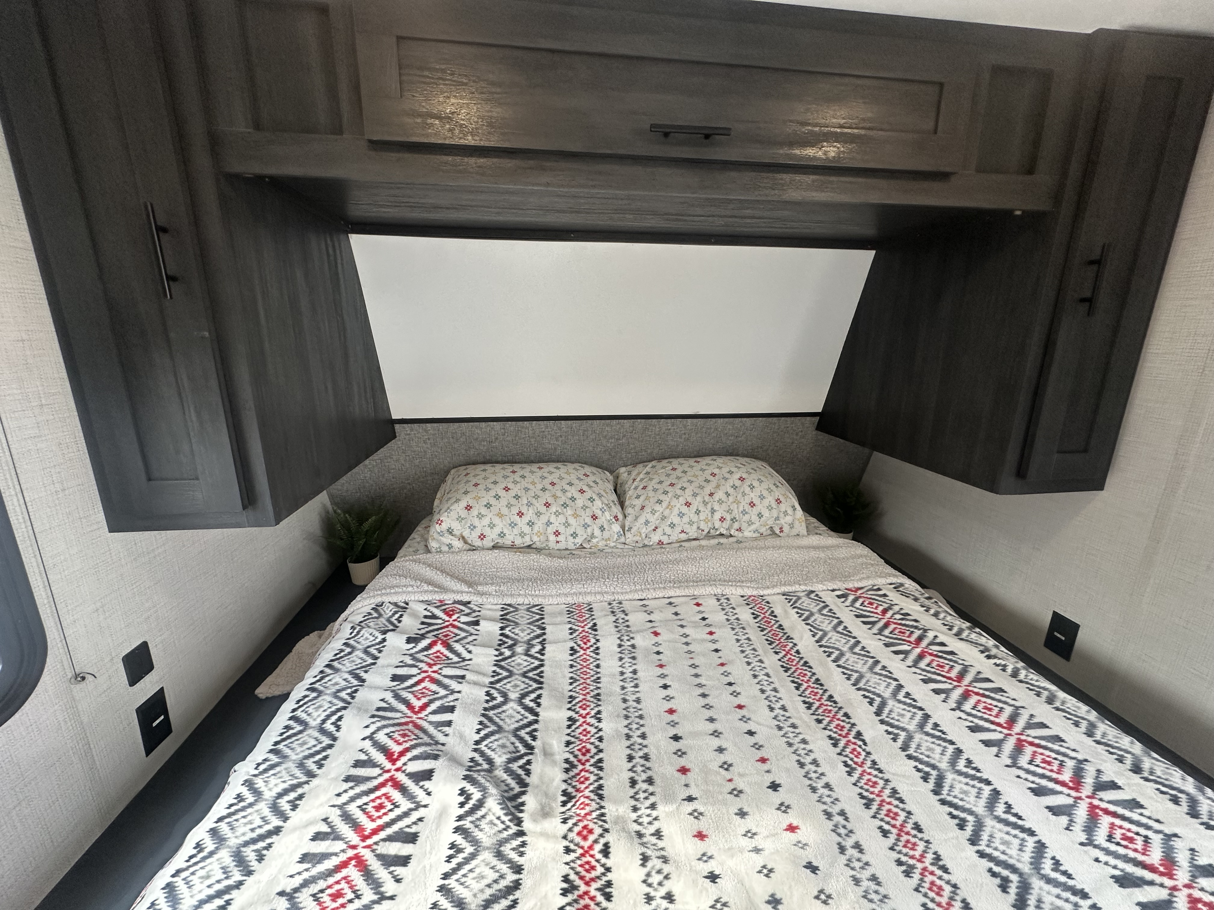 This cozy RV rental bedroom features a comfy bed with a patterned blanket, overhead storage, and integrated outlets.. Ozark Lite 2021