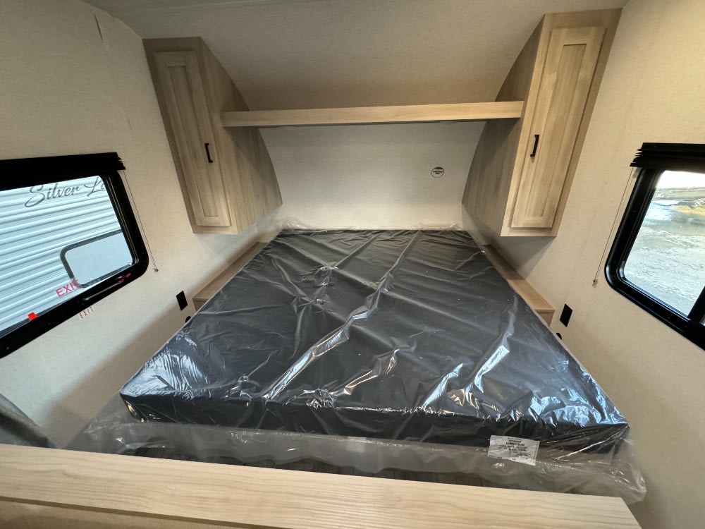 This RV rental bedroom features a new bed with protective plastic, overhead wood cabinets, and two windows.. Forest River East toWest SilverLake 25bhle 2024