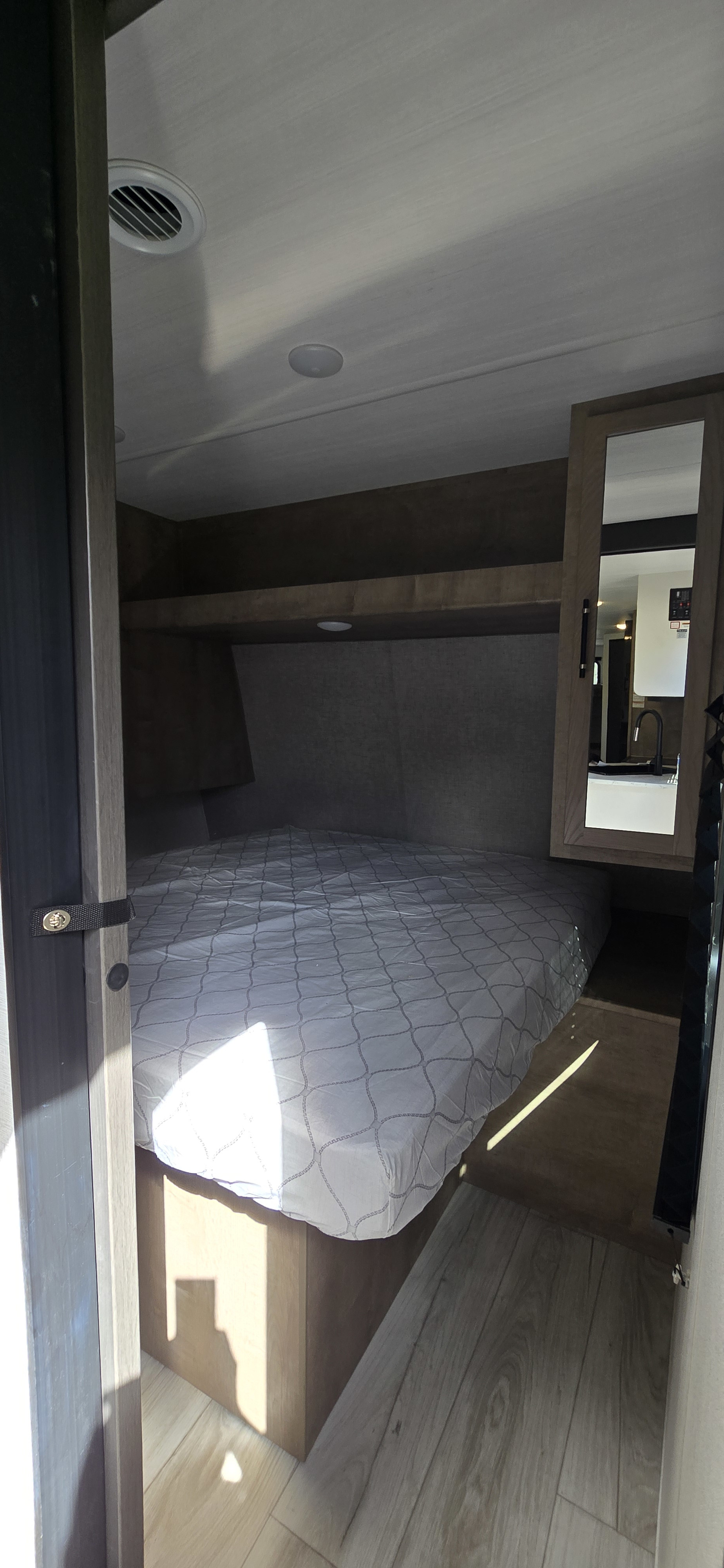 Inside a bright RV rental: a bedroom with a patterned mattress, overhead storage, and a mirrored cabinet reflecting the kitchen.. Keystone Springdale 2025