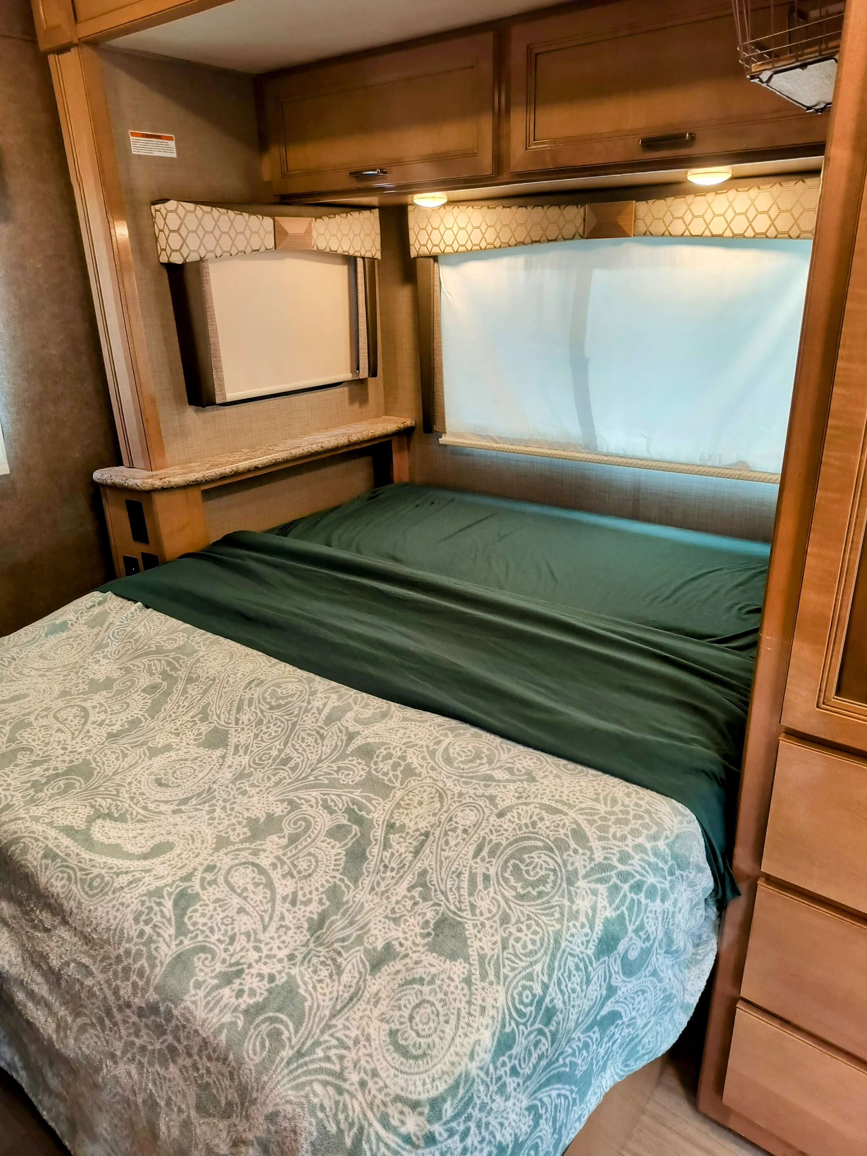 RV rental bedroom with a bed featuring green and paisley bedding, overhead cabinets, and window blinds.. Thor Motor Coach Quantum 2020