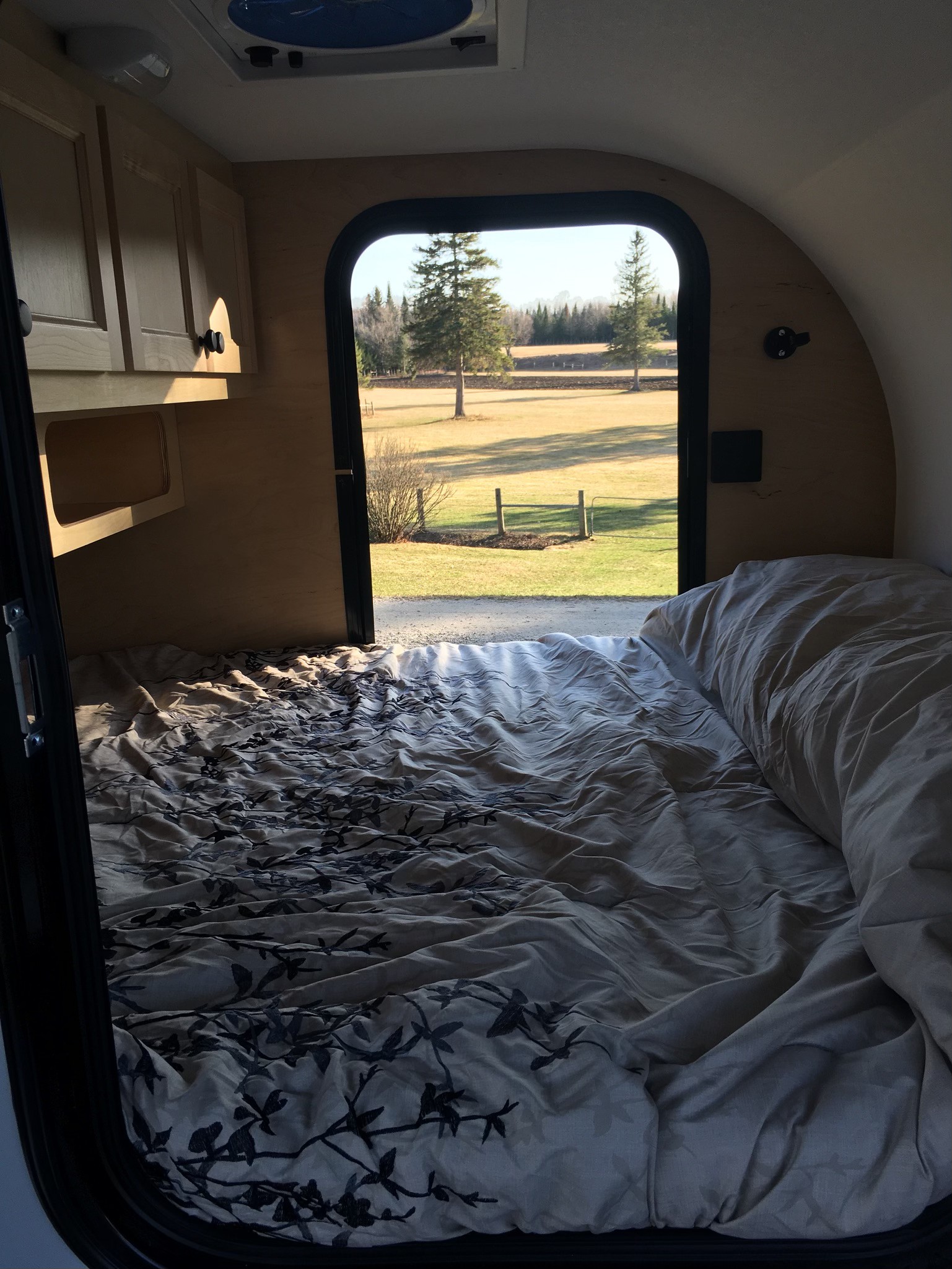 Cozy RV rental interior: bed with patterned duvet, open door reveals sunny field, trees, nature.. Car-go Trailers Liberty2go XL 2021