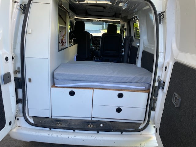 Inside a white camper van RV rental: compact bed, under-bed storage drawers, and a side cabinet. Ready for adventure!. NISSAN NV200 2015