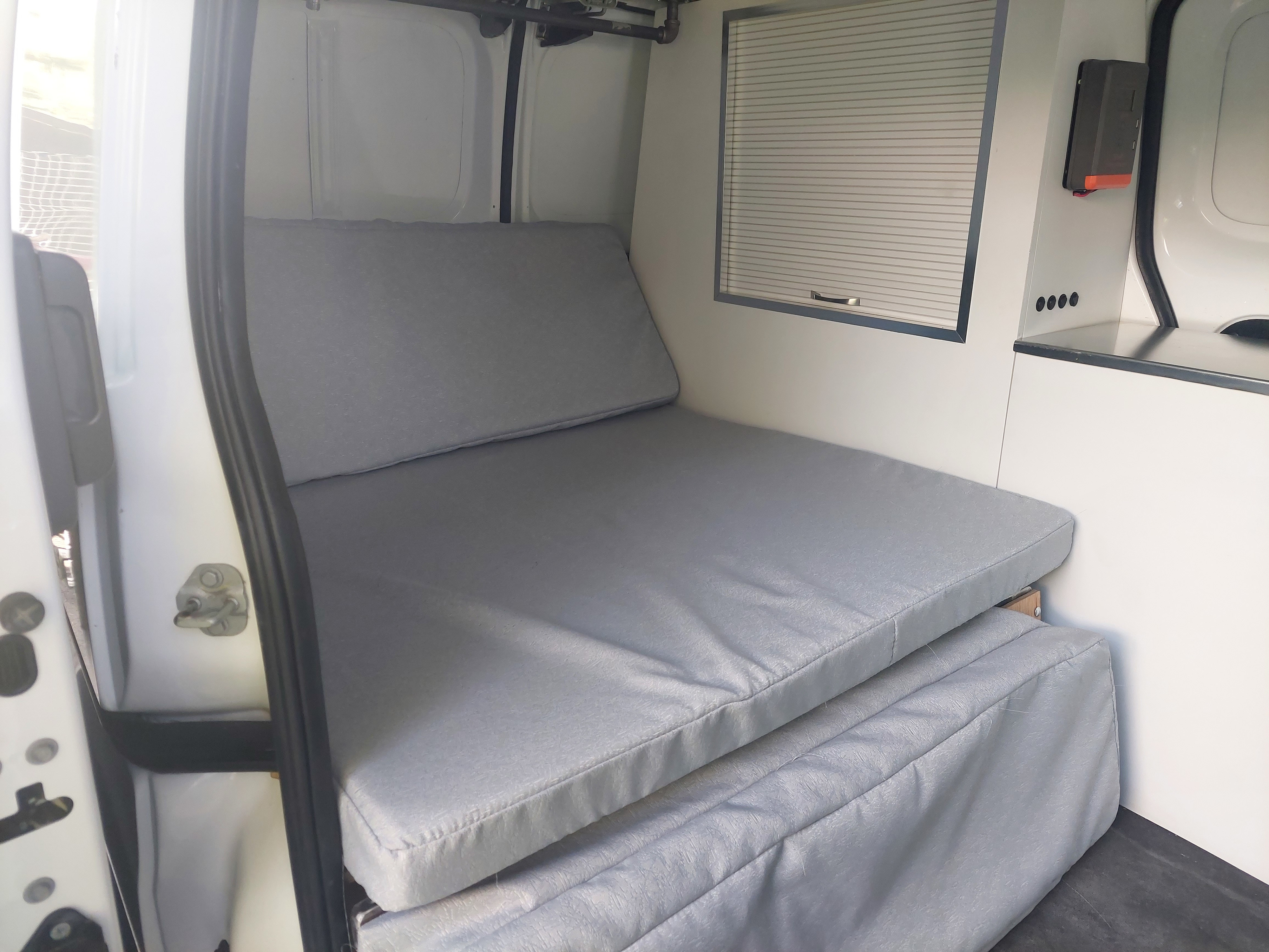 This **RV rental** features a comfortable grey folding bed, custom interior cabinets, and a window blind for privacy.. NISSAN NV200 2015