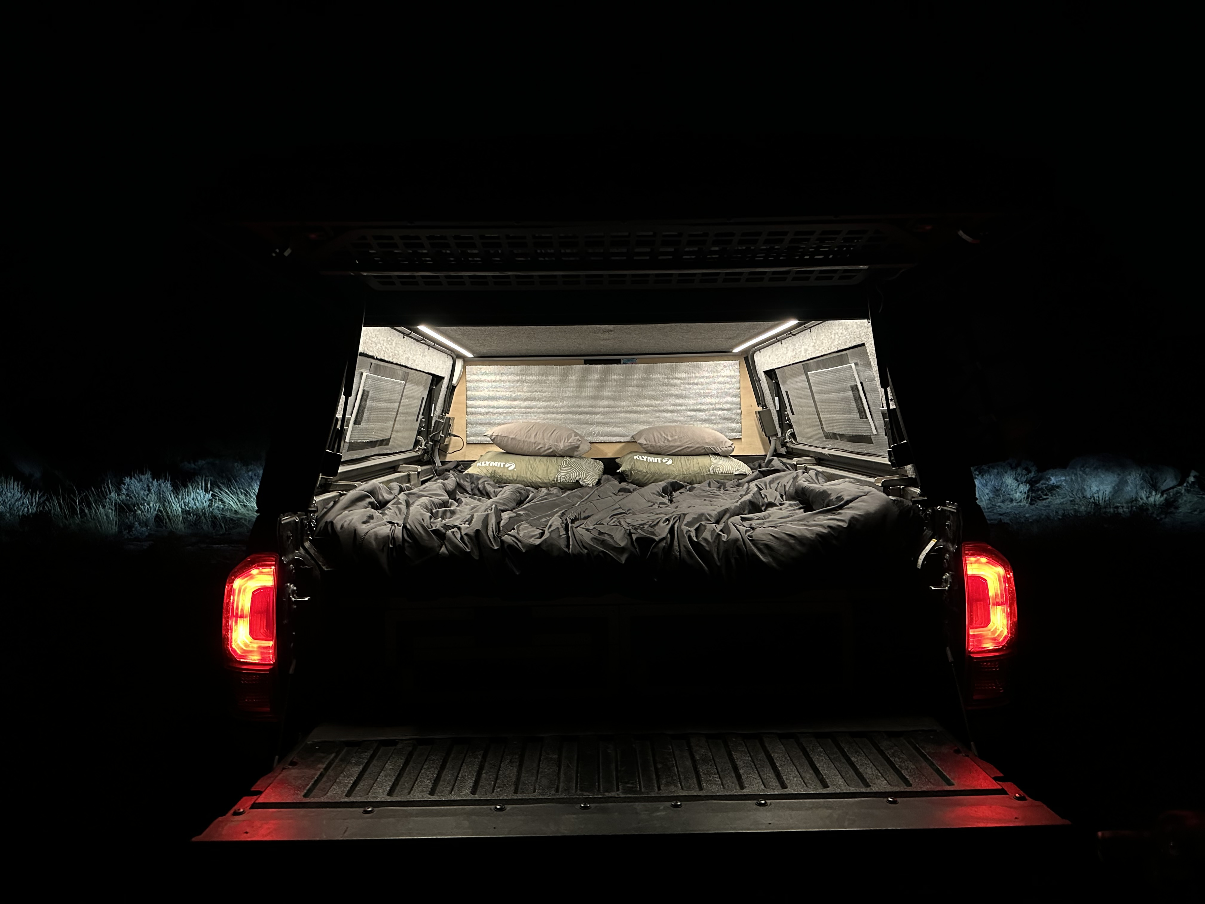 Cozy truck bed camping setup at night for an RV rental. Features illuminated bed, open tailgate, and glowing red taillights.. Alu-Cab Toyota Tacoma 2023