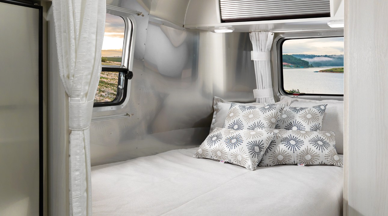 Relax in this RV rental: a comfortable bed with patterned pillows and iconic windows revealing diverse scenic views.. Airstream Bambi 2022