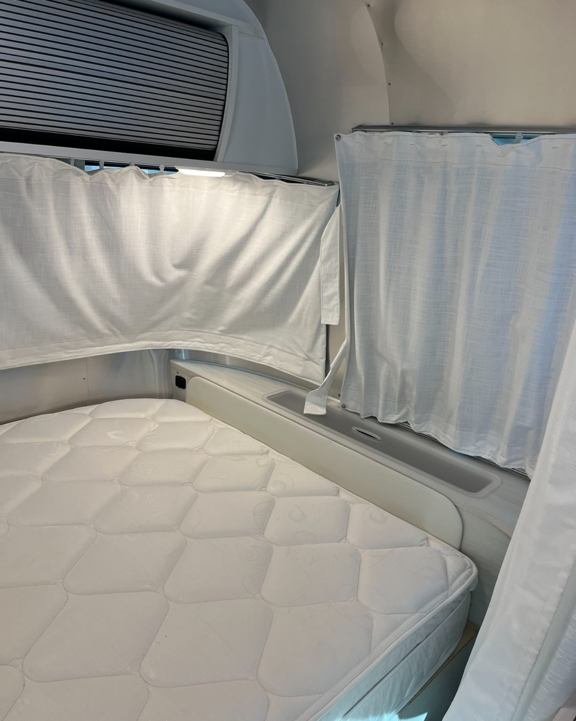 Clean RV rental interior showing a comfortable white mattress, curtains, and sleek bedroom area for your next trip.. Airstream Bambi 2021