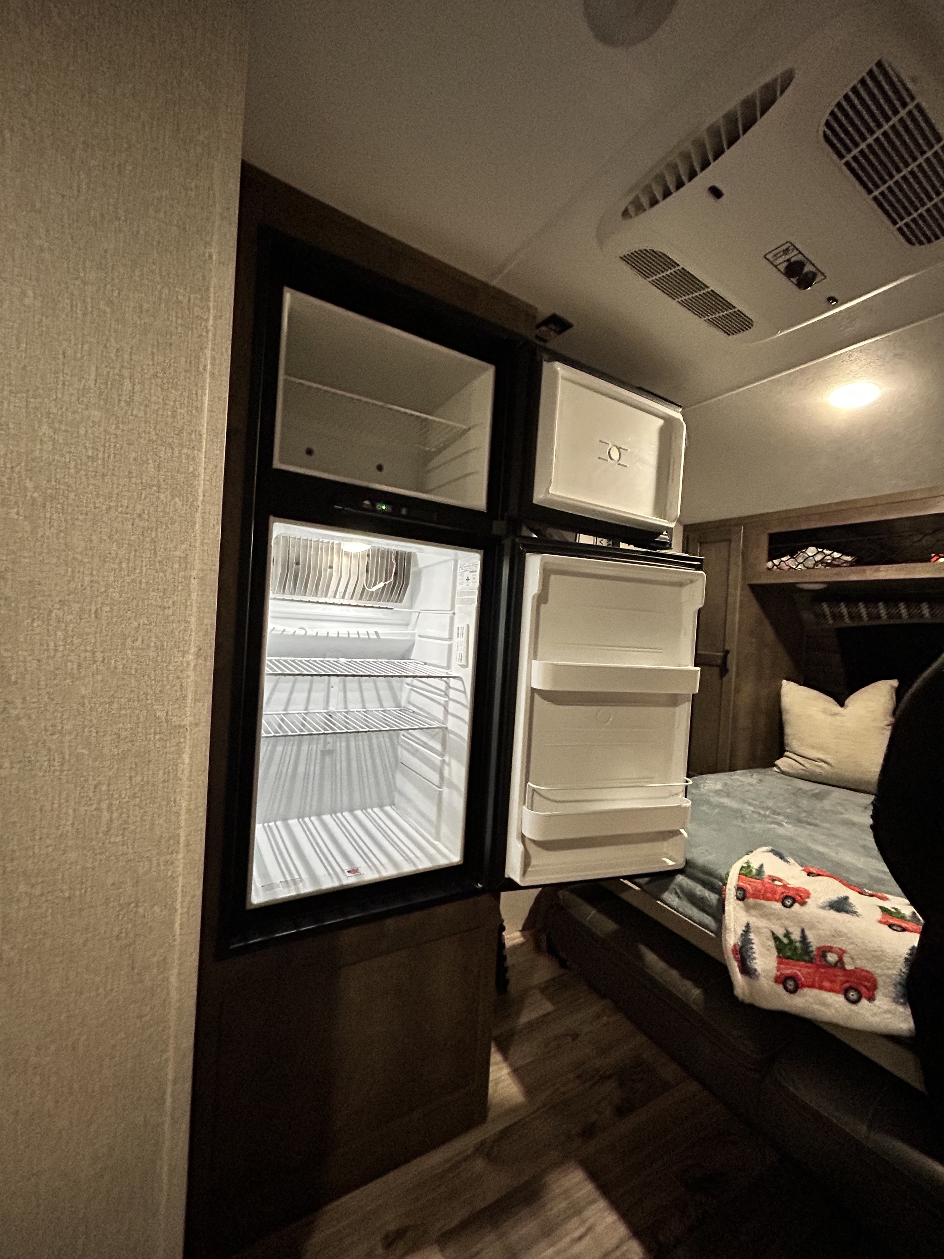 RV rental interior featuring an open compact refrigerator, revealing its shelves. A bed with a festive blanket is visible in the background.. Venture SONIC LITE 2022