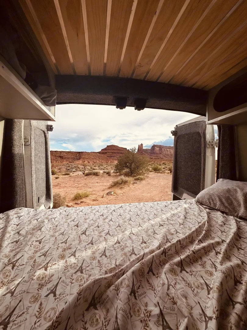 Inside an RV rental, a cozy bed with an Eiffel Tower sheet faces a breathtaking desert panorama of red rock mesas and a tree.. FORD TRANSIT 2017