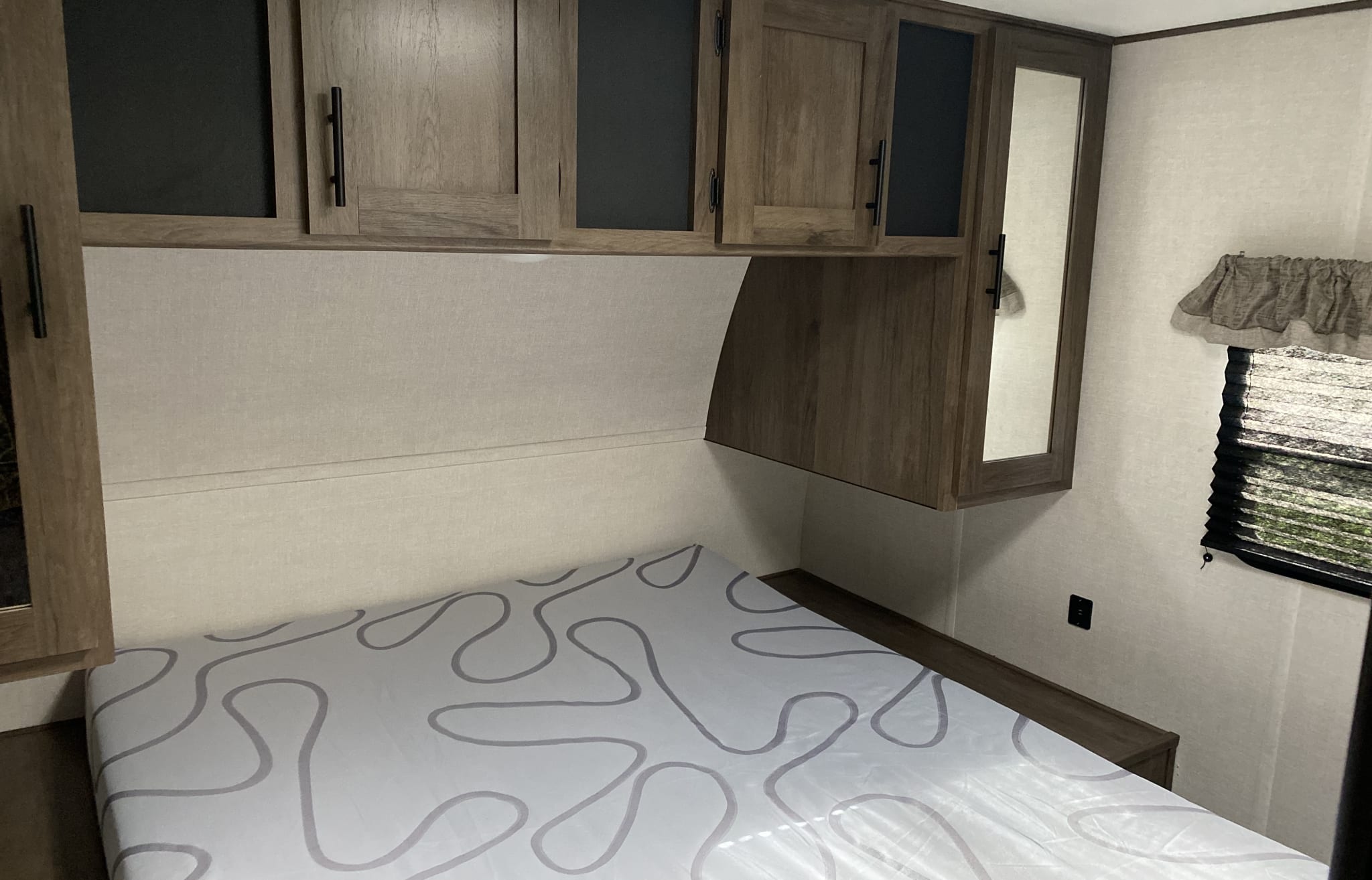 An RV rental bedroom with a patterned mattress, wooden overhead storage cabinets, and a side window with blinds.. Prime Time Avenger 2021