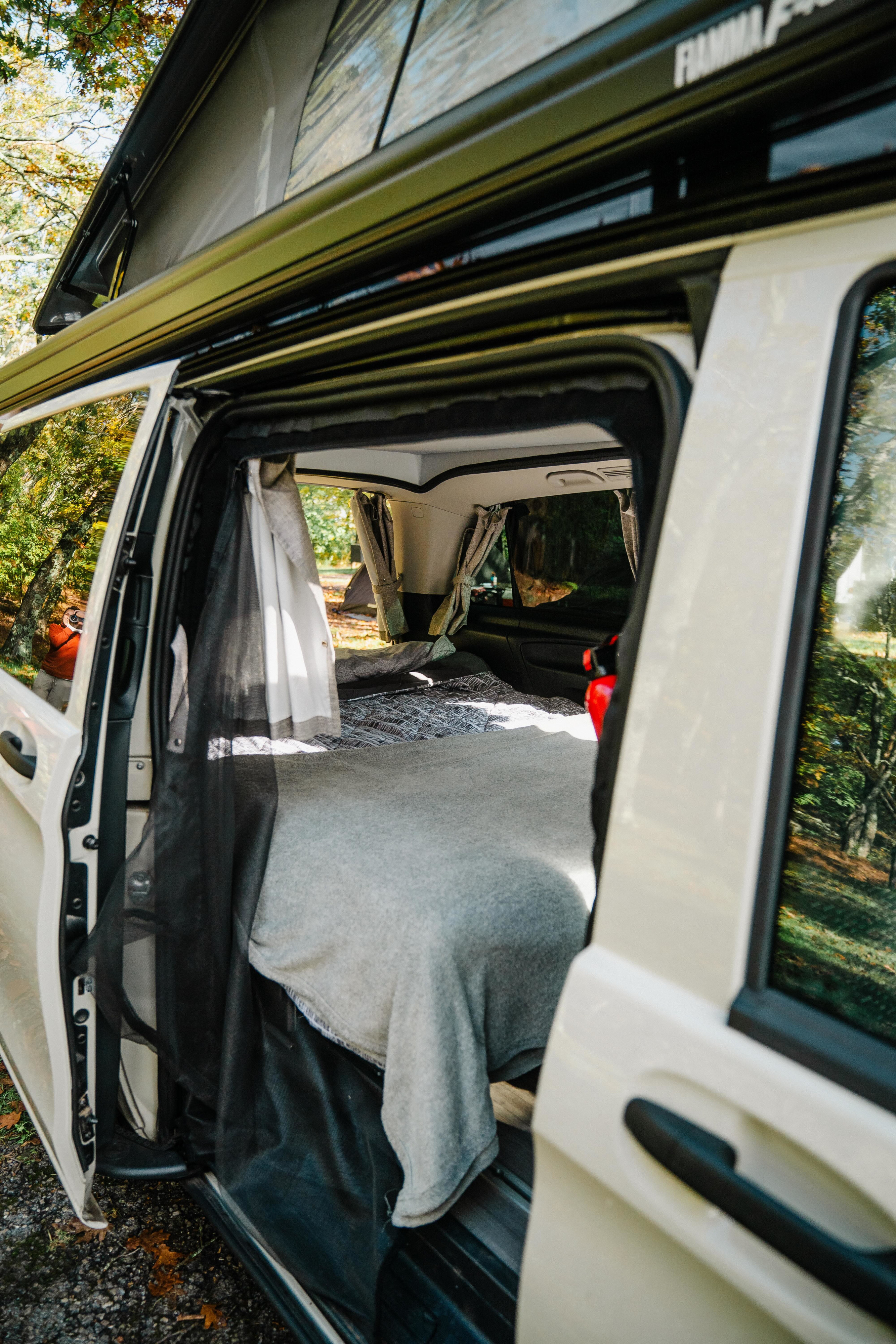 Open door of an RV rental camper van reveals a cozy bed, pop-top roof, and mesh screen for comfortable outdoor stays.. Mercedes Metris 2021