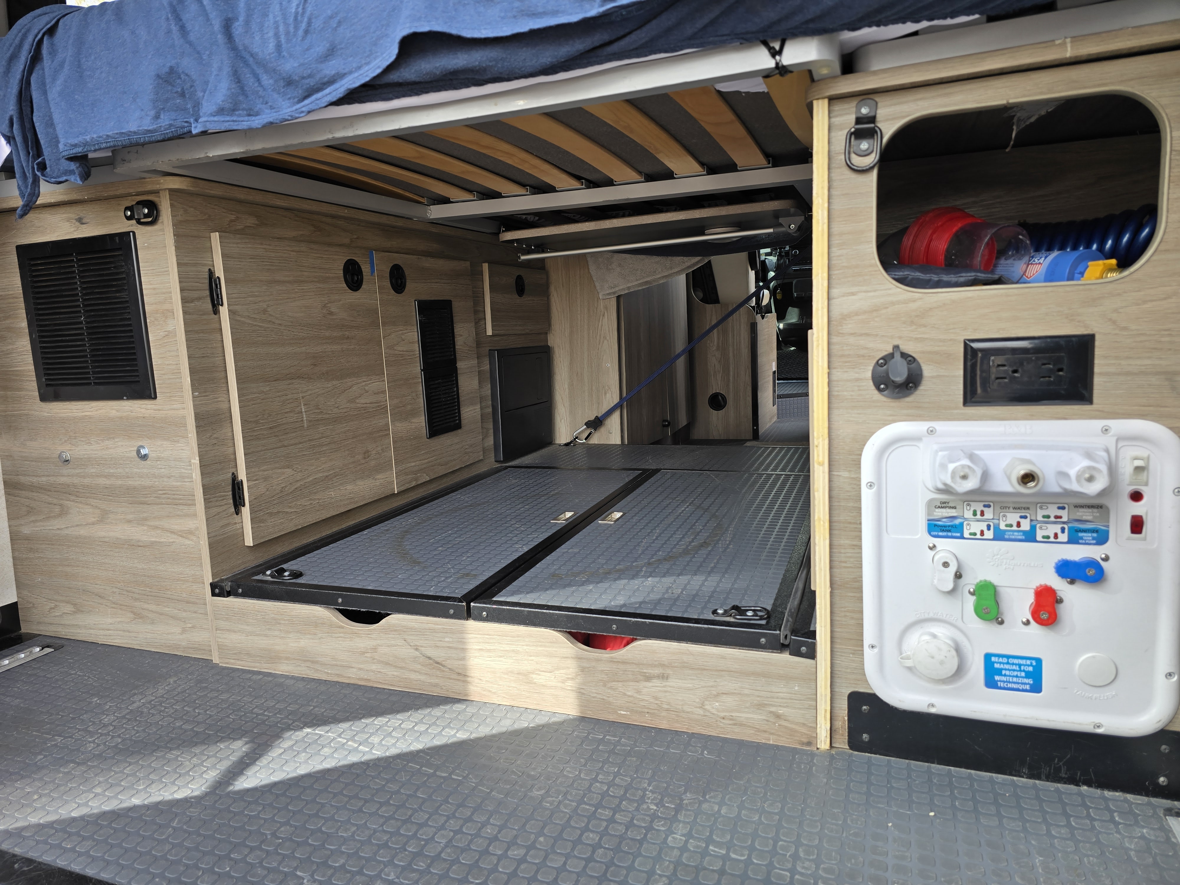 RV rental's rear cargo area shows pull-out storage, water utility panel, and bed frame for organized adventures.. Winnebago Solis 2023
