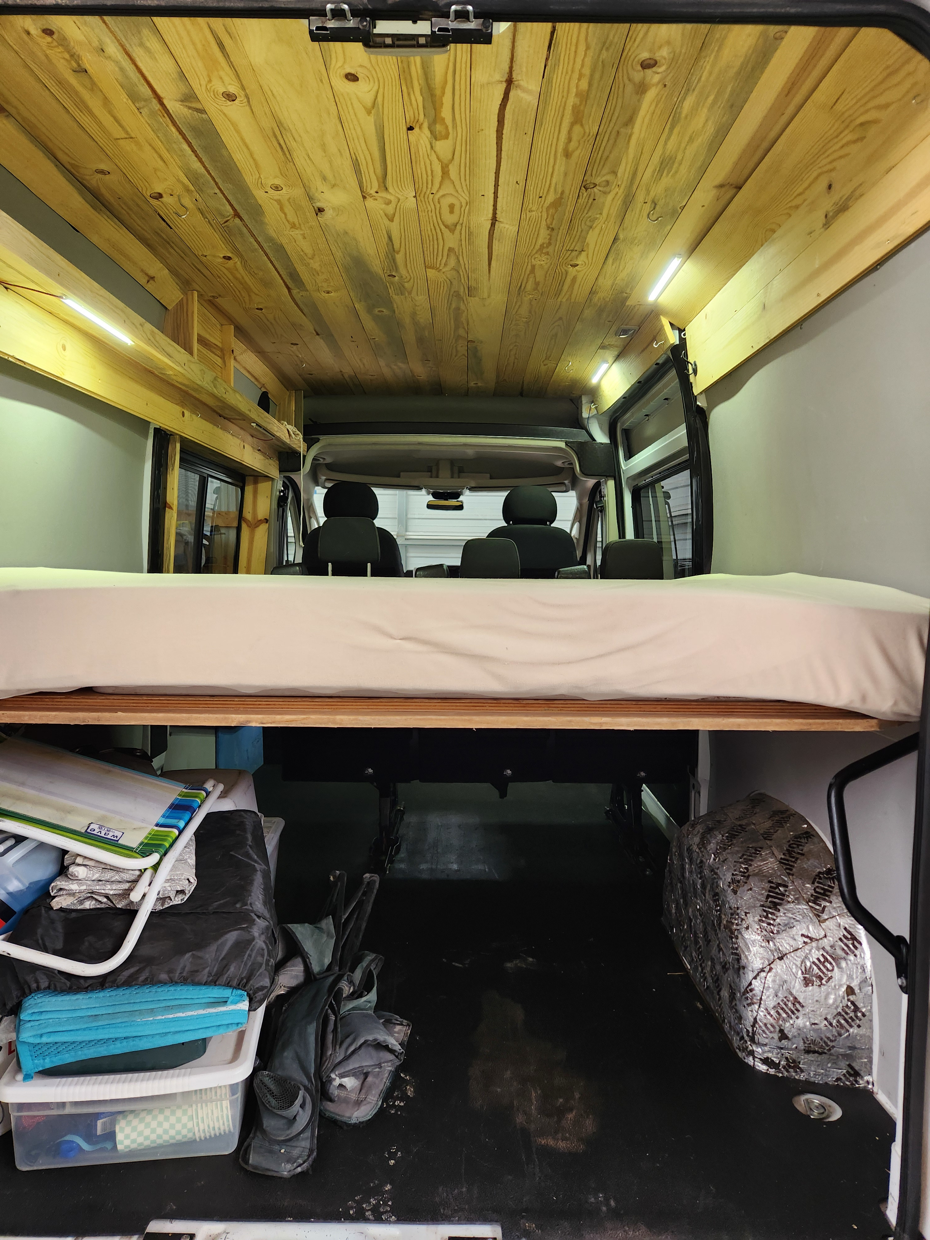 Inside an RV rental conversion: A wooden-paneled ceiling with lights, a comfortable bed, and under-bed storage are visible, facing the front cab.. Ram Promaster 2020