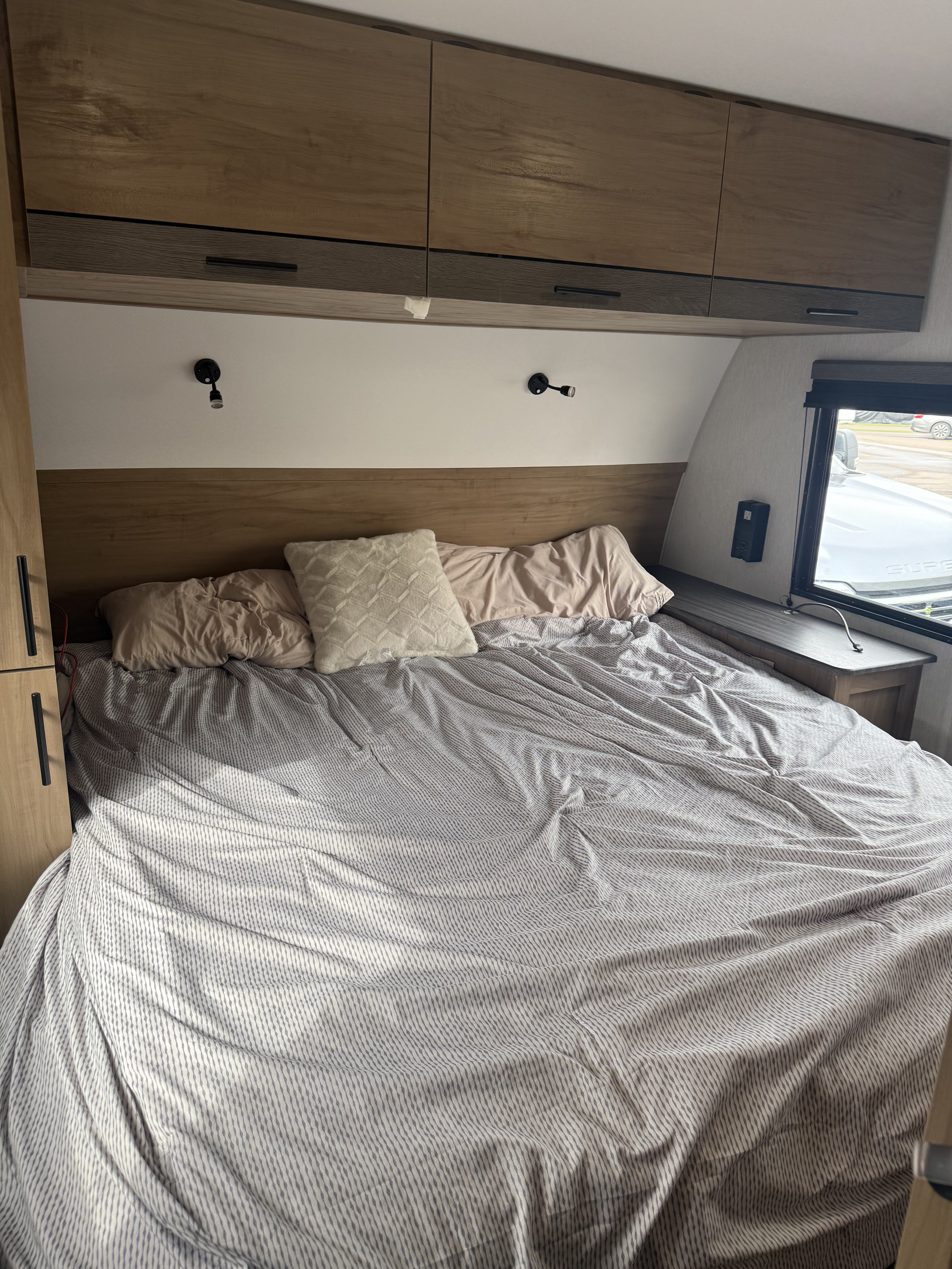 Cozy bedroom in an RV rental, featuring a striped bed, pillows, overhead cabinets, and a side window.. Forest River Campsite Reserve 23CC 2024