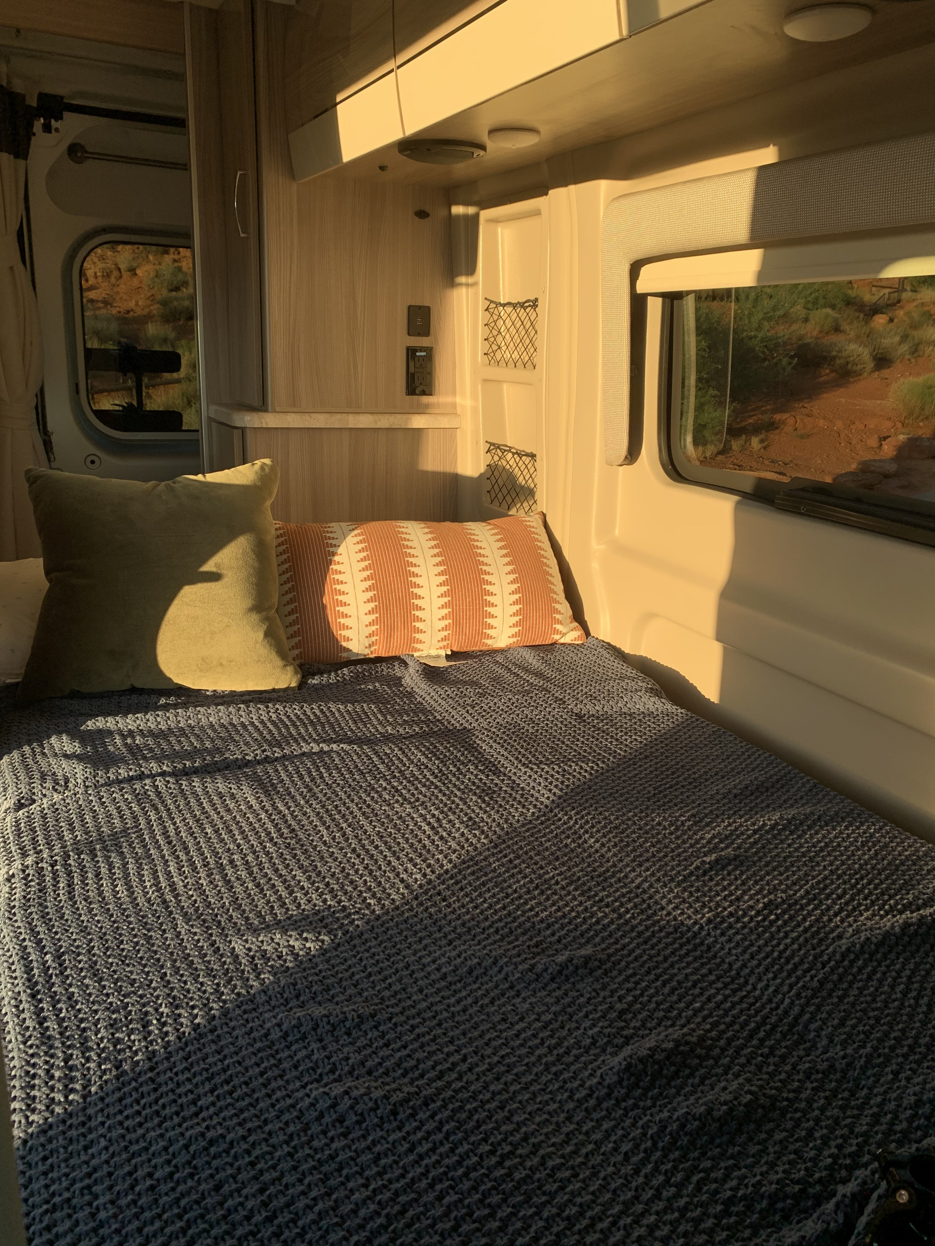 Inside an RV rental: a sunlit bed with blue blanket and pillows, framed by windows offering desert landscape views.. Thor Motor Coach Scope 2023