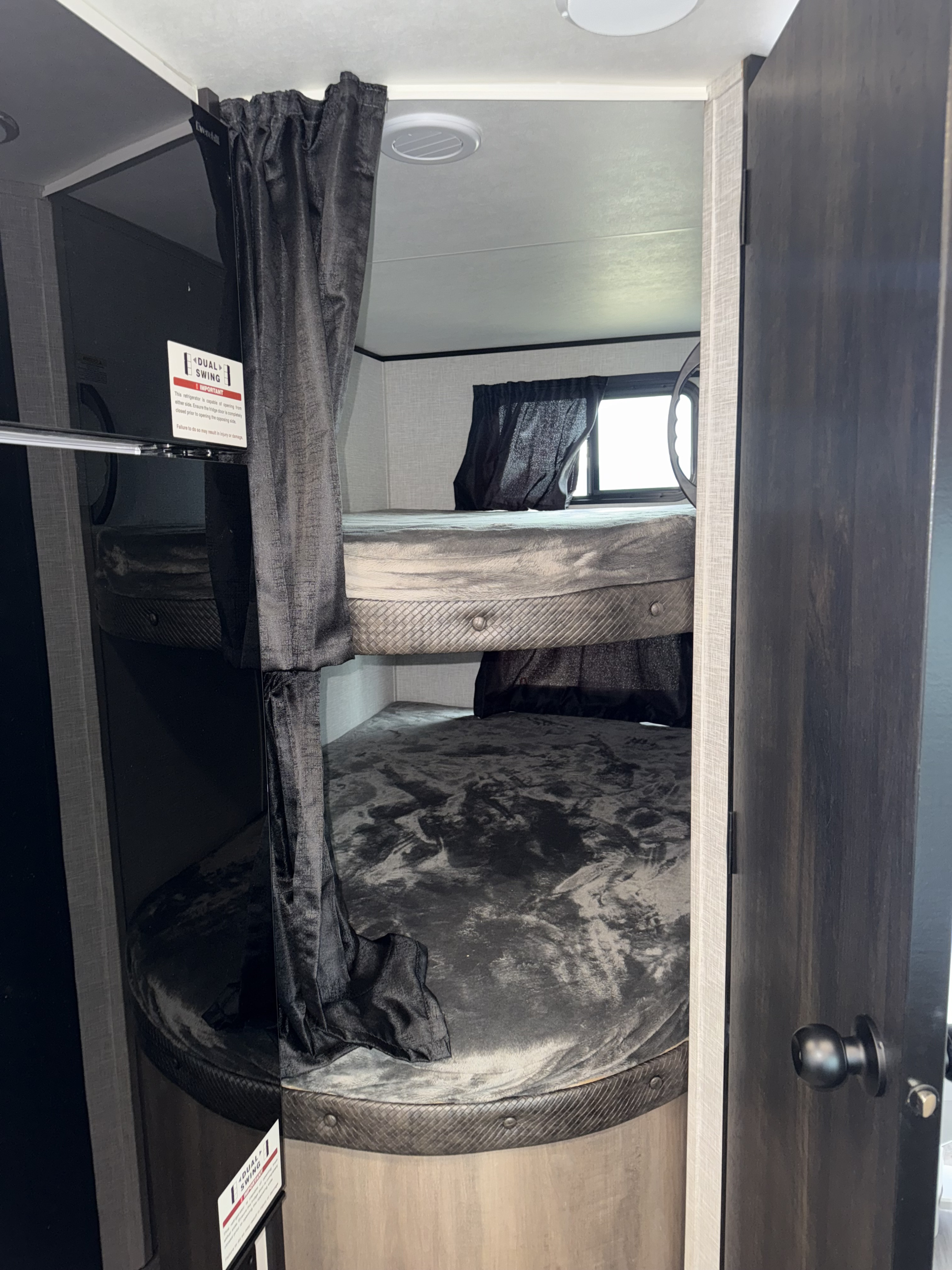 Inside an RV rental: two bunk beds with dark curtains, next to a black dual-swing Everchill refrigerator.. Jayco Jay Flight 2024