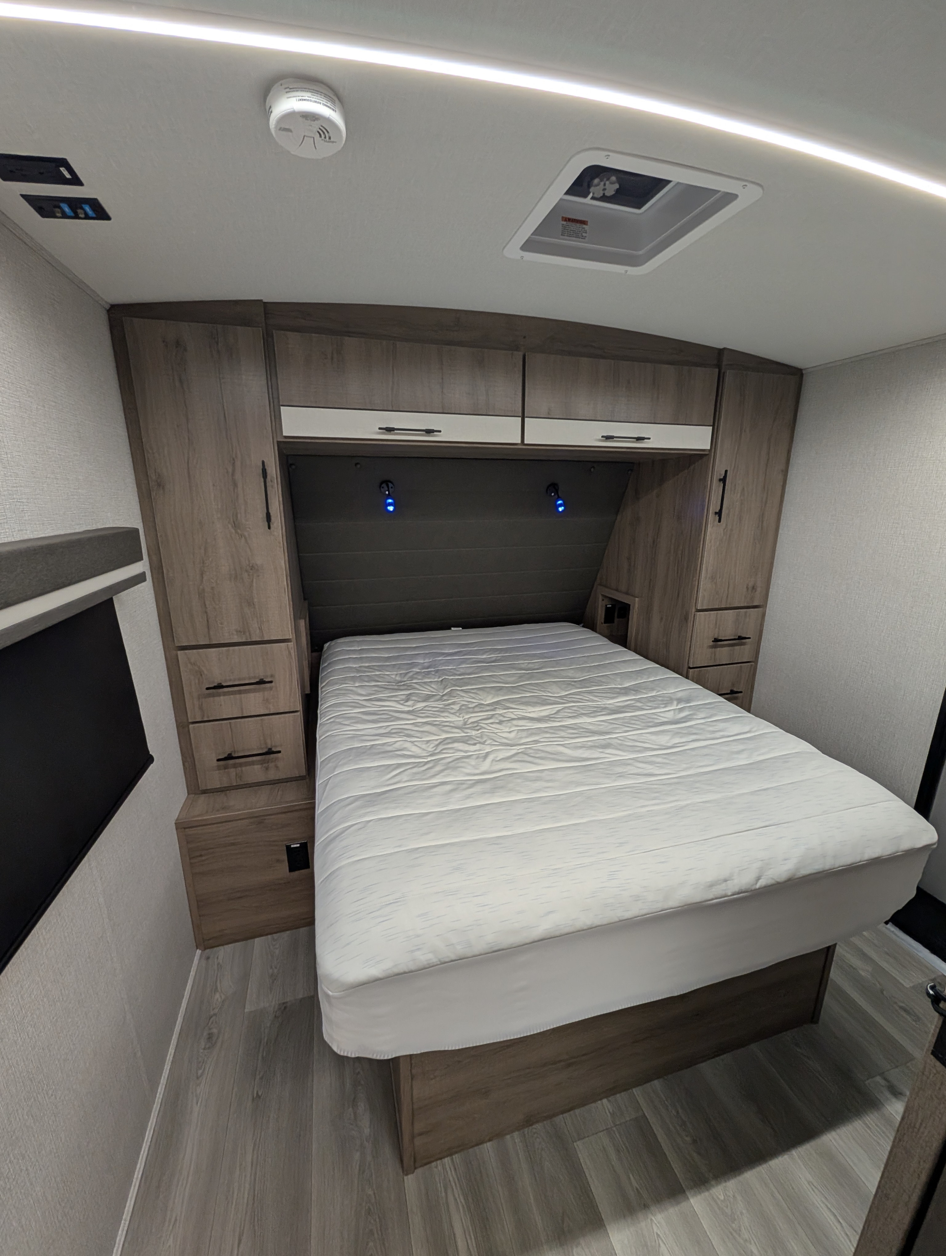 Modern RV rental bedroom: features a white bed, overhead cabinets, side storage, ceiling vent, and linear lighting.. Grand Design Imagine 2024