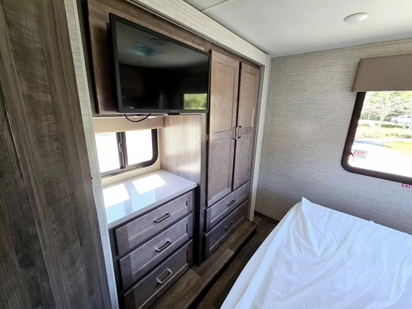 RV rental bedroom: TV, wood wardrobe, dresser, and a bed with white sheets next to a window showing an outdoor view.. Winnebago Minnie Winnie 2024