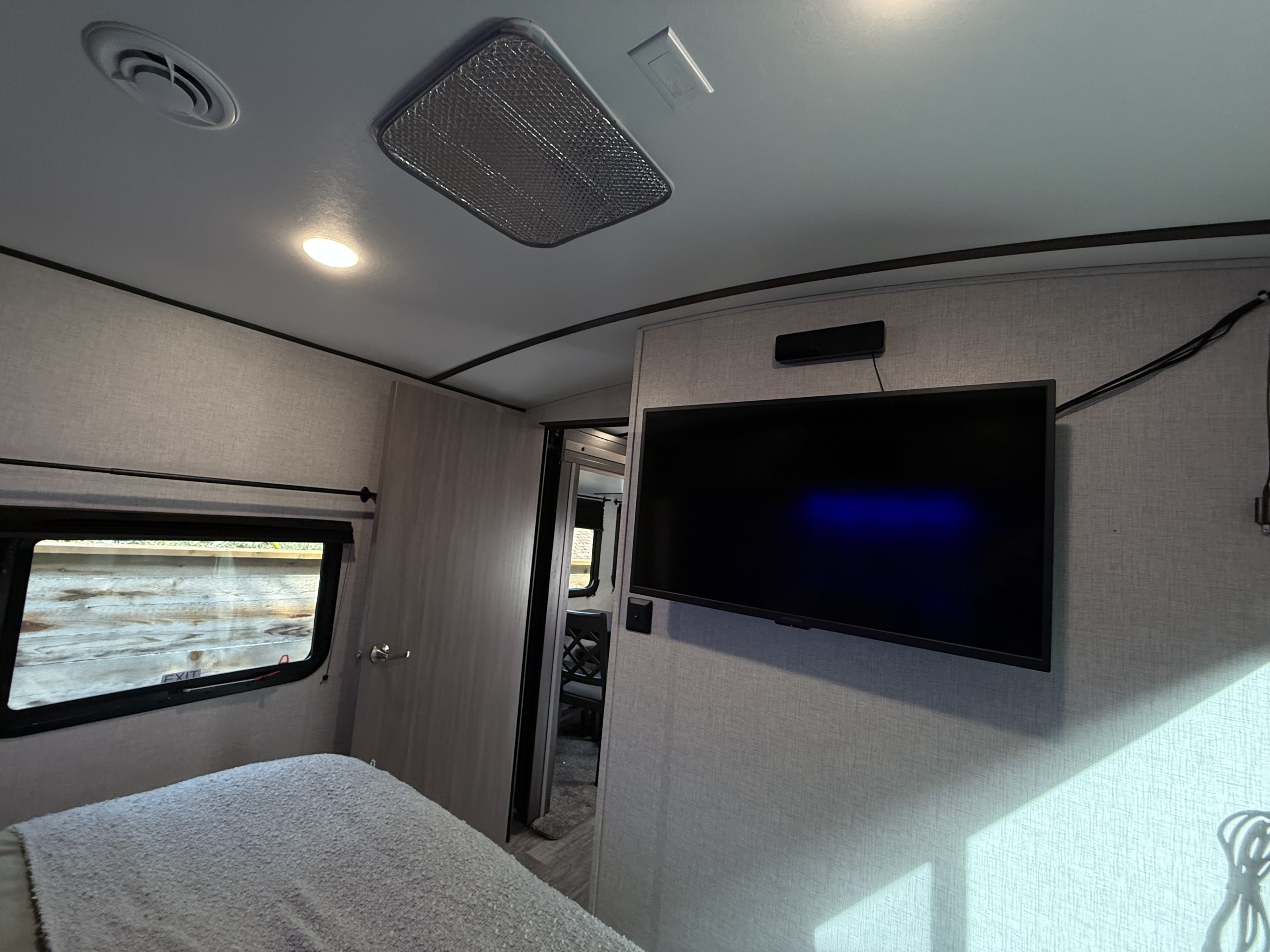 Inside a compact RV rental bedroom, featuring a bed, window with "EXIT" sign, TV, and ceiling AC vent and light, creating a cozy space.. Crossroads Sunset Trail Super Lite 2021