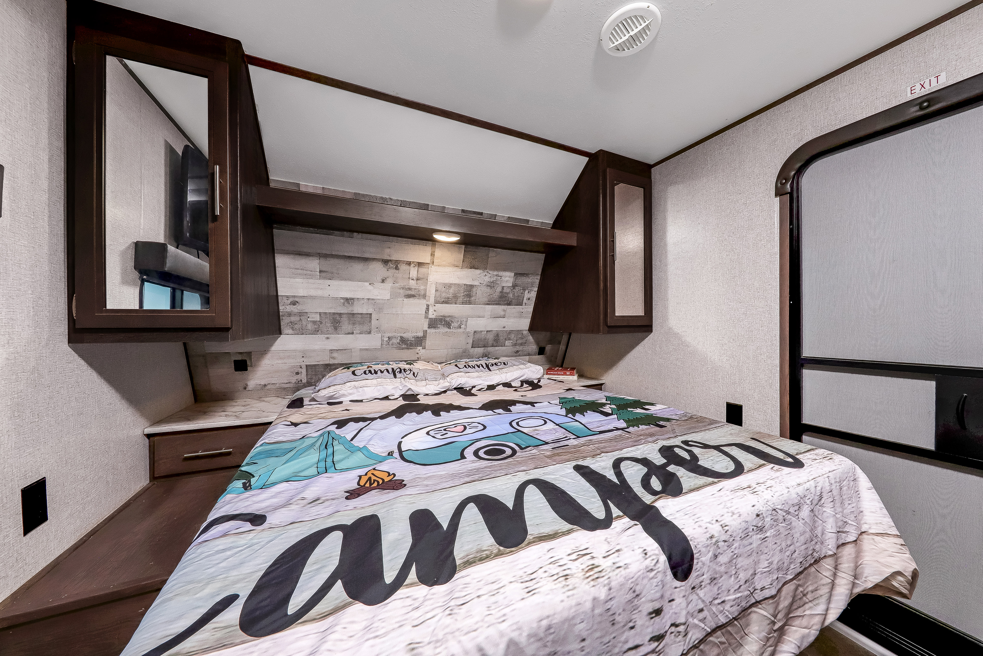 Cozy RV rental bedroom with fun 'Camper' themed bedding, overhead storage, and a nearby exit door.. Dutchmen Coleman 2021
