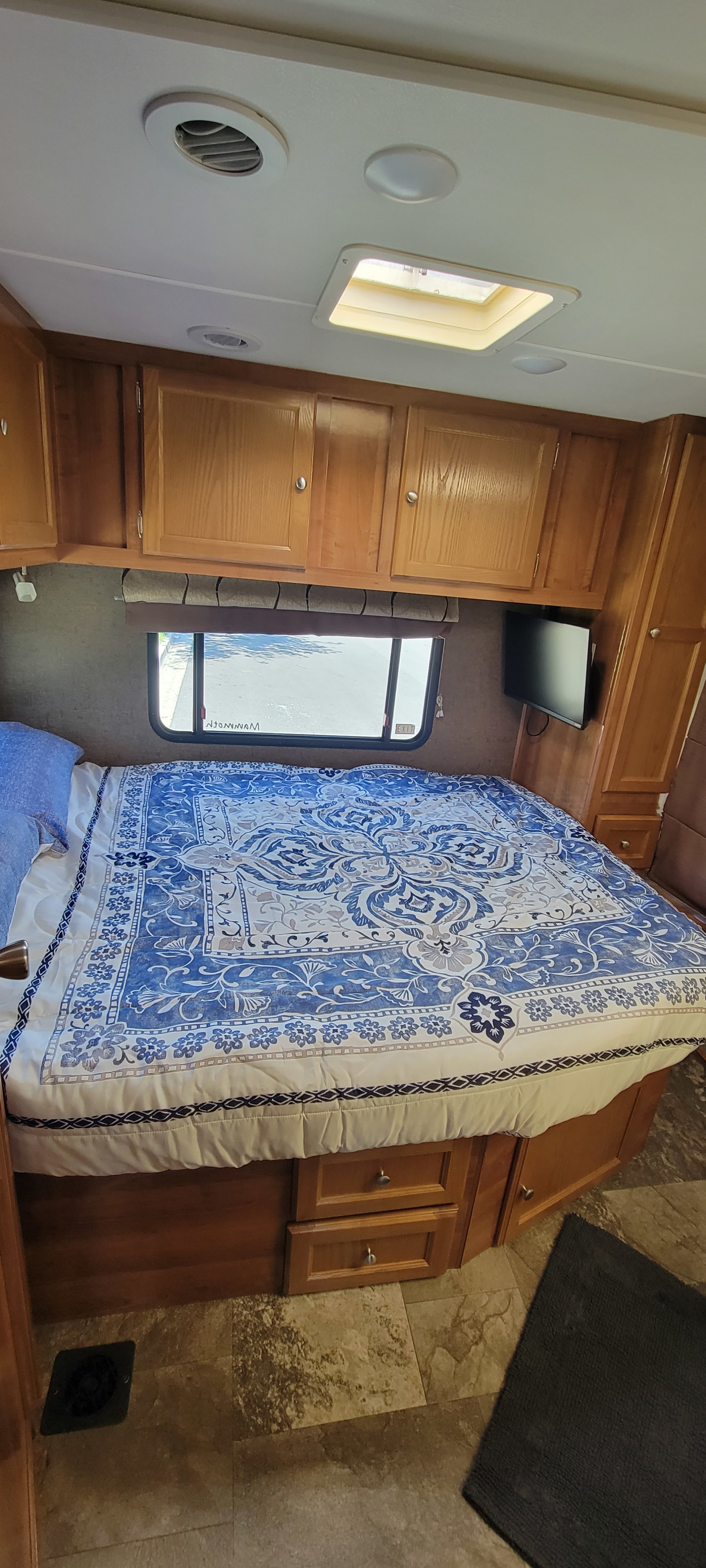Cozy RV rental bedroom with a large bed, blue patterned comforter, overhead wood cabinets, window, and TV.. Forest River Coachmen Leprechaun 2018
