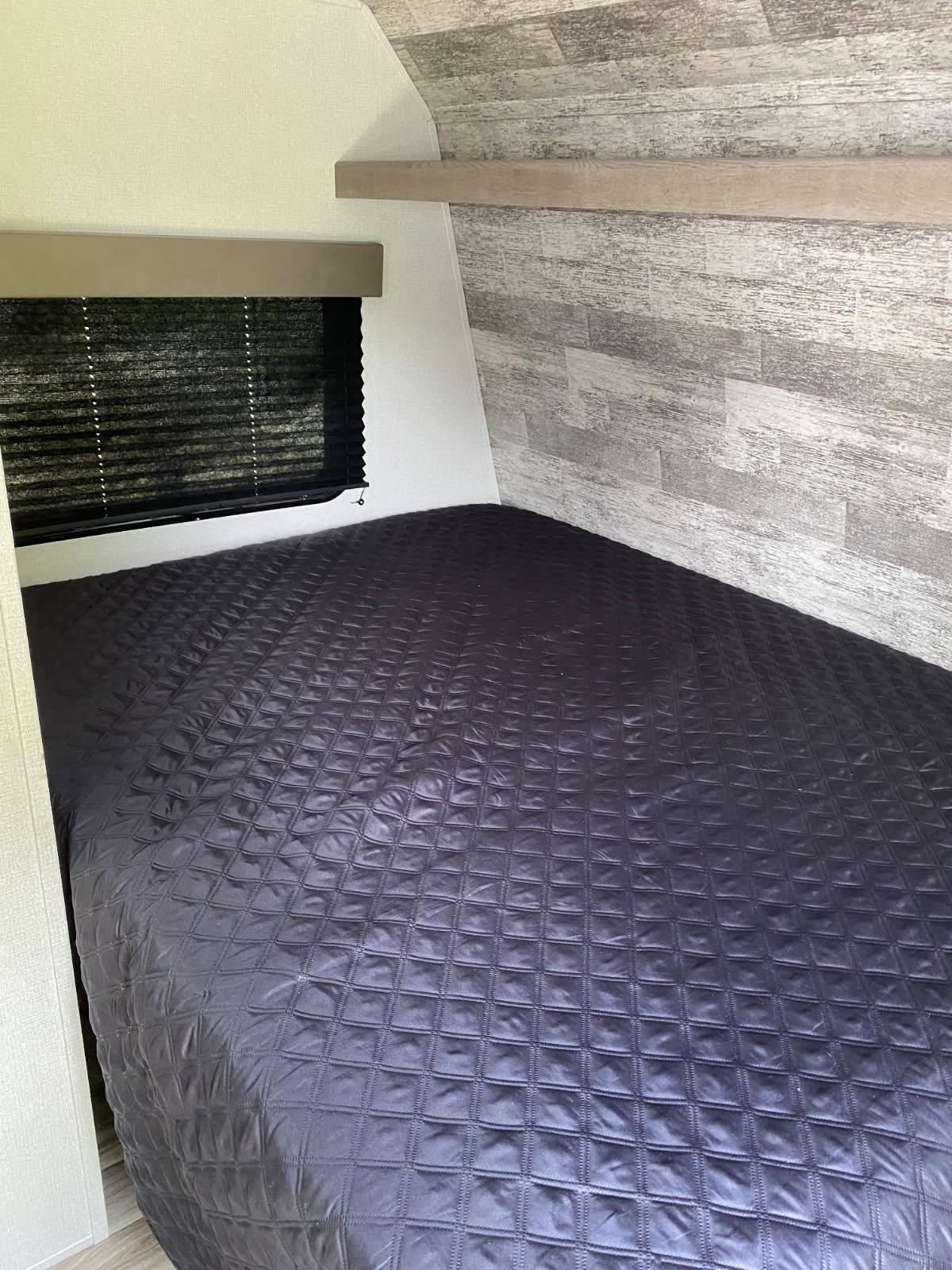 Cozy RV rental bedroom featuring a dark, quilted bed, window with blinds, and modern wood-look wall paneling.. Keystone RV Springdale 2021