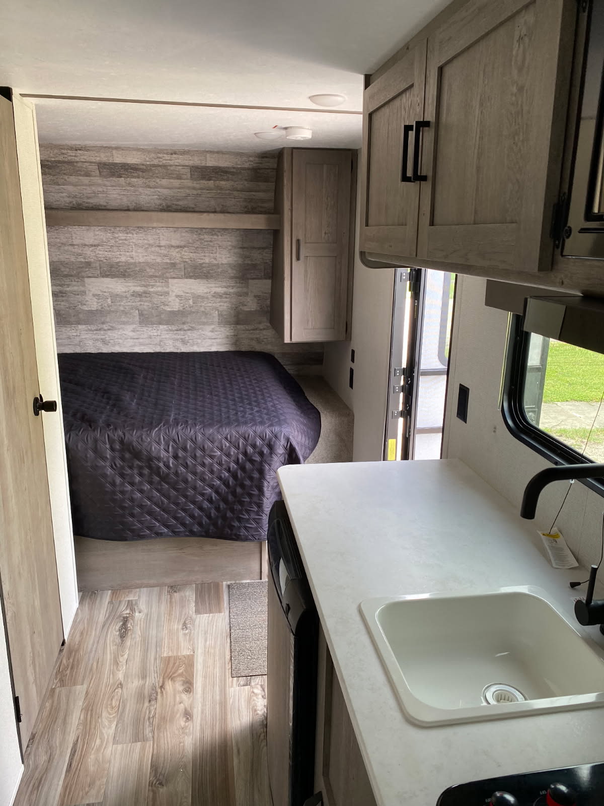 RV rental interior: cozy bedroom with dark bedspread and wood-look wall, plus a kitchen with white counter and sink.. Keystone RV Springdale 2021