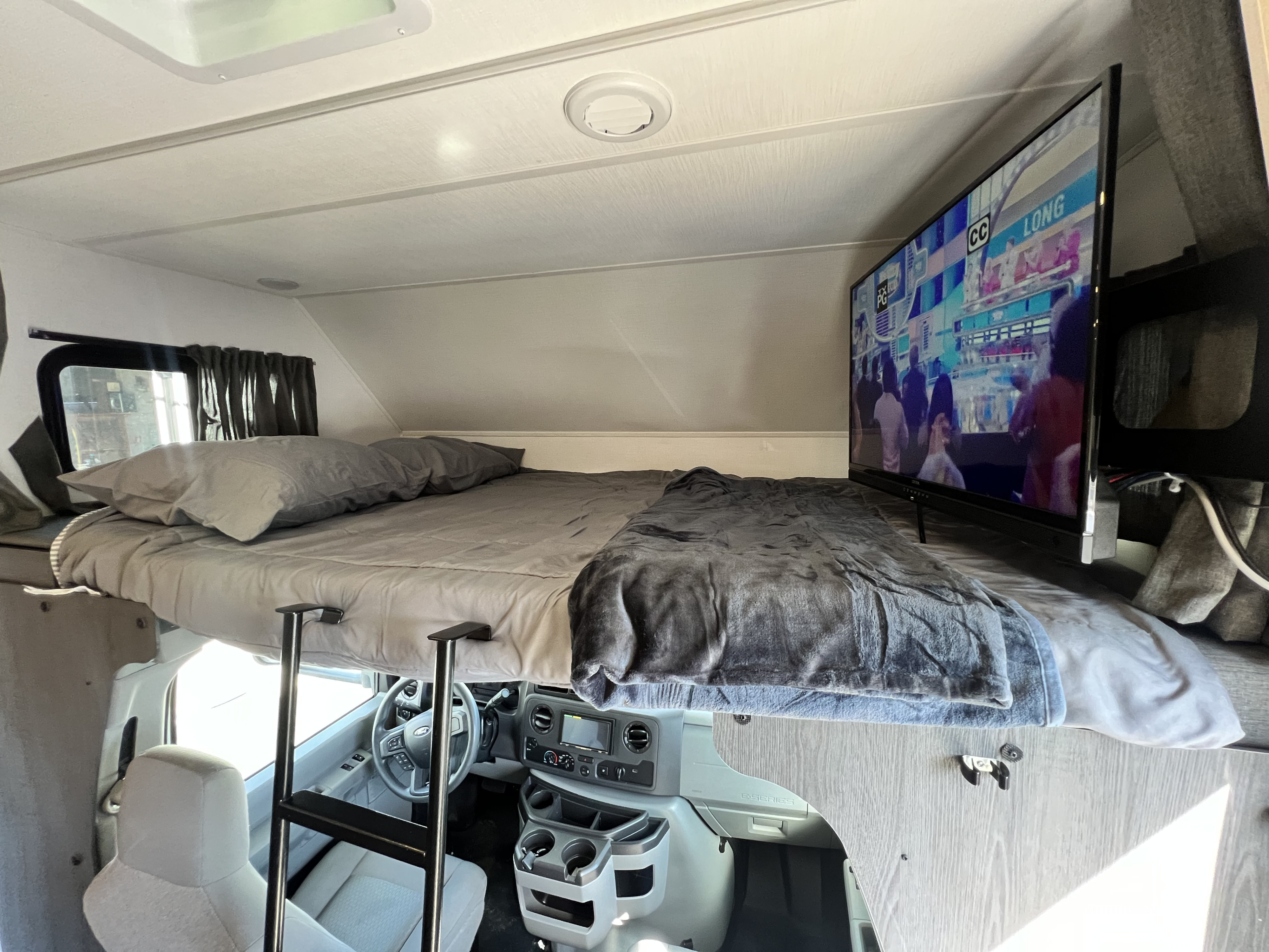 View inside an RV rental: a comfortable cab-over bunk bed with TV, accessible by ladder, overlooking the vehicle's dashboard.. Forest River Sunseeker 2024