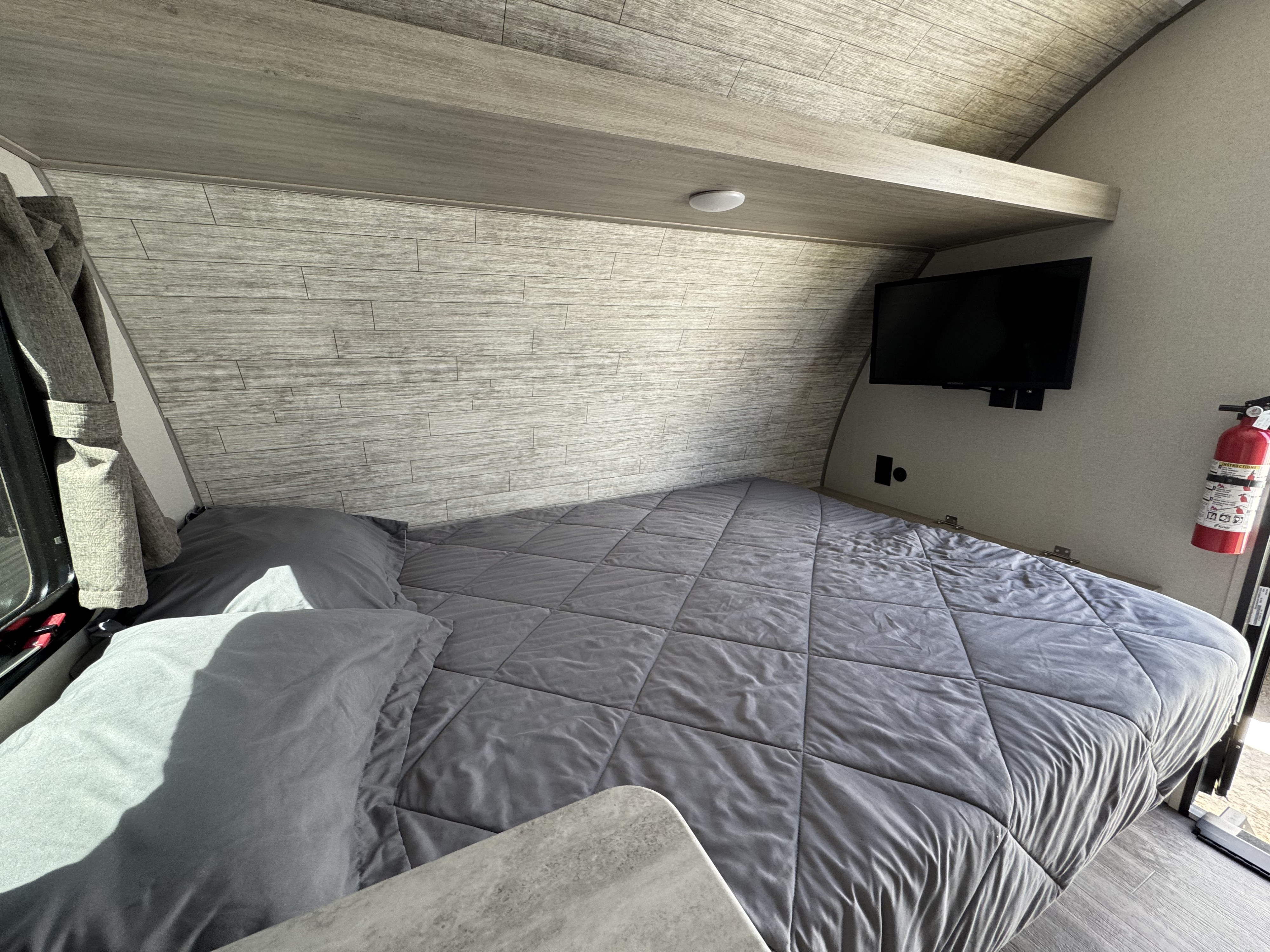 Cozy RV rental bedroom featuring a grey bed, overhead shelf, and wall-mounted TV. Perfect for road trip comfort.. Forest River Aurora 13B 2025