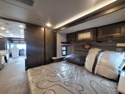 Modern RV rental bedroom with a full bed, plastic-covered bedding, and overhead storage cabinets, showing a spacious interior.. Forest River Coachmen Sportscoach SRS 2023