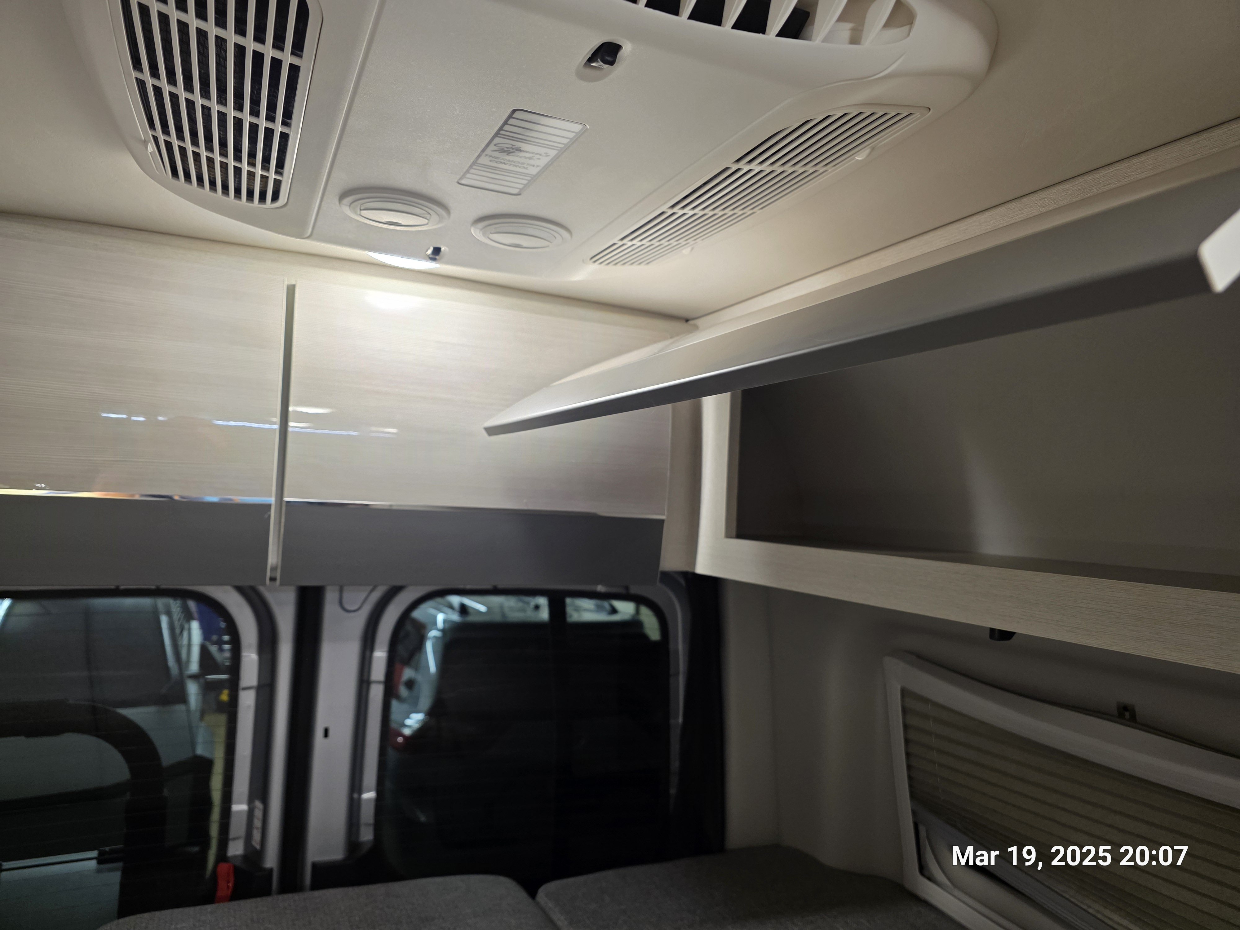 Modern RV rental interior featuring overhead AC vents, lighting, and light-toned storage cabinets above tinted windows.. Thor Tranquility 19M 2025