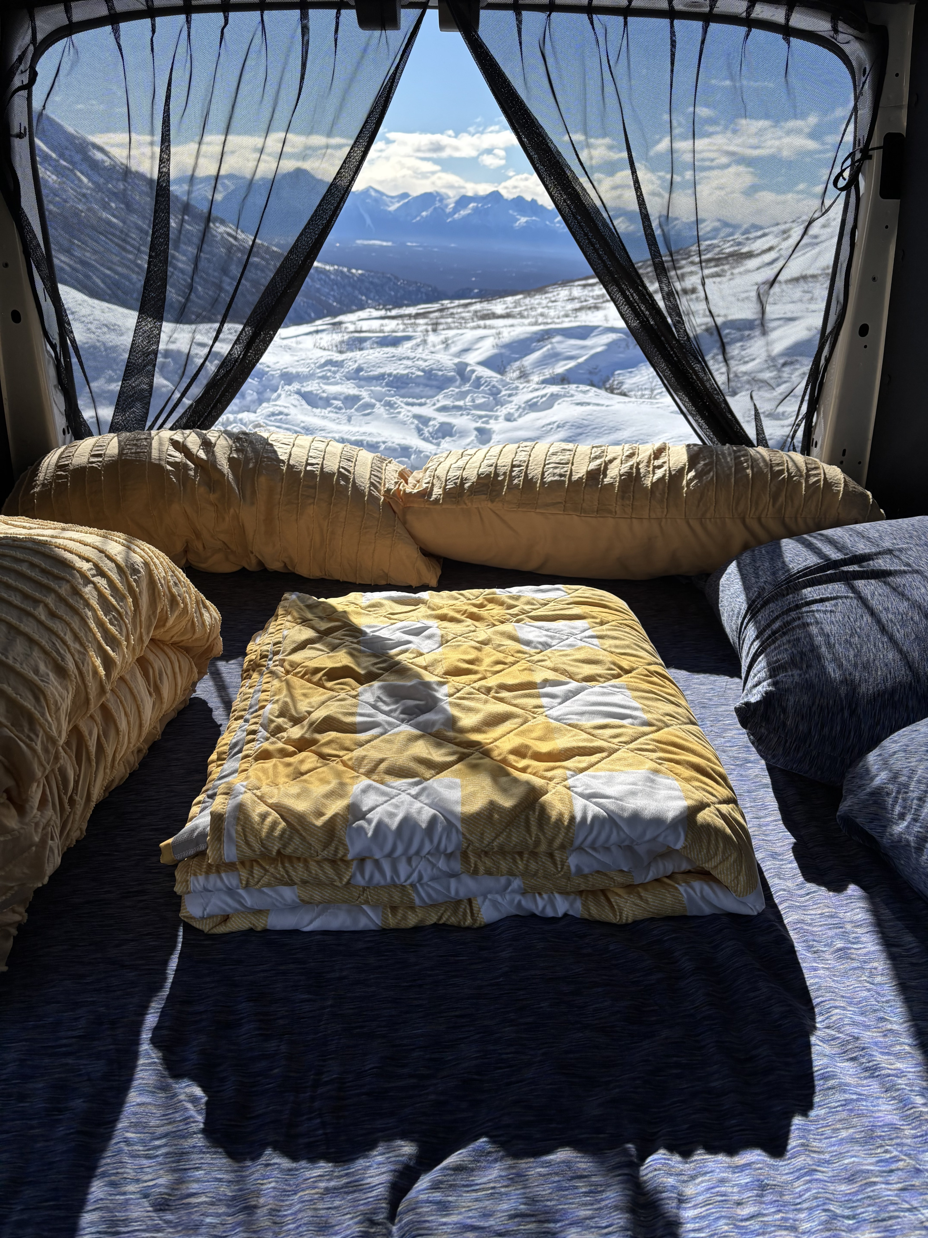An RV rental's cozy interior, with a soft bed and pillows, offers breathtaking snowy mountain views.. Ford Transit 2023