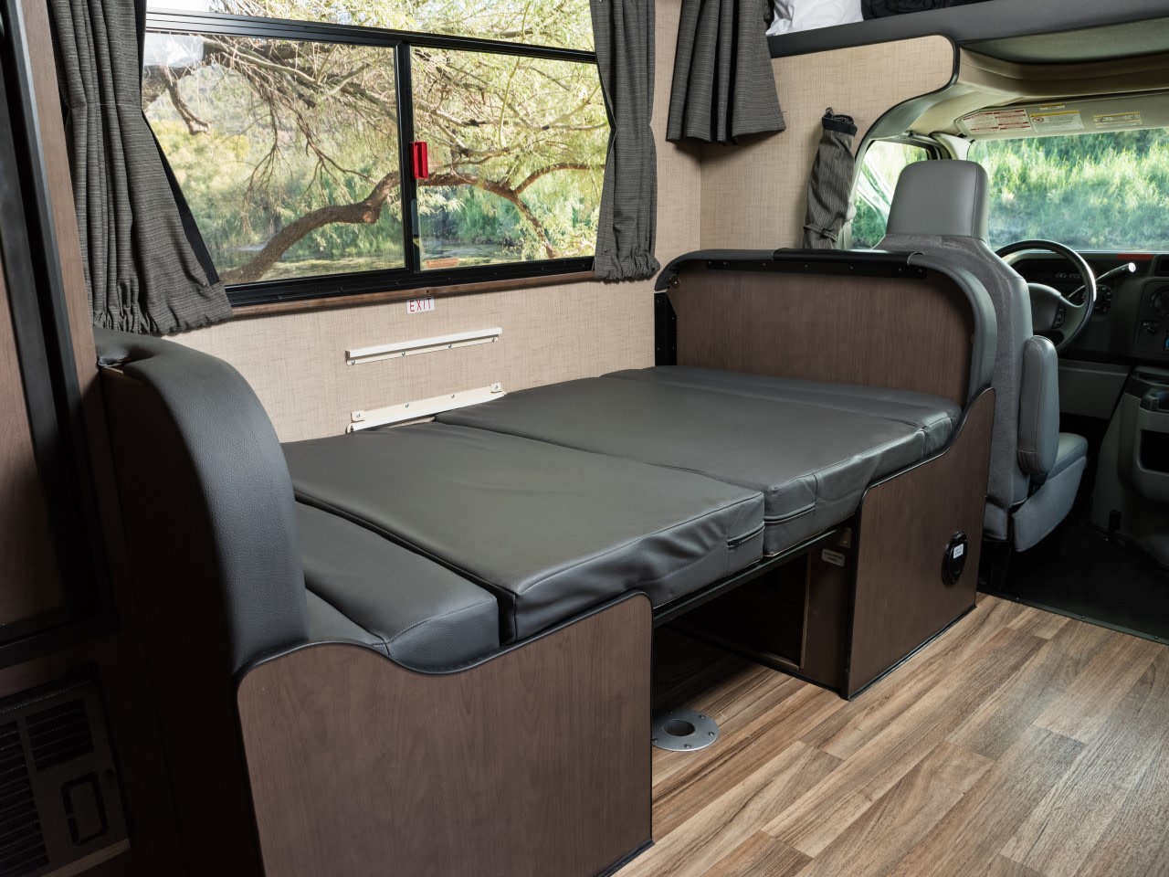 RV rental interior: Grey convertible bed/sofa by a window, offering a tranquil view. Ideal for travel & relaxation.. Thor Ford E350 2020