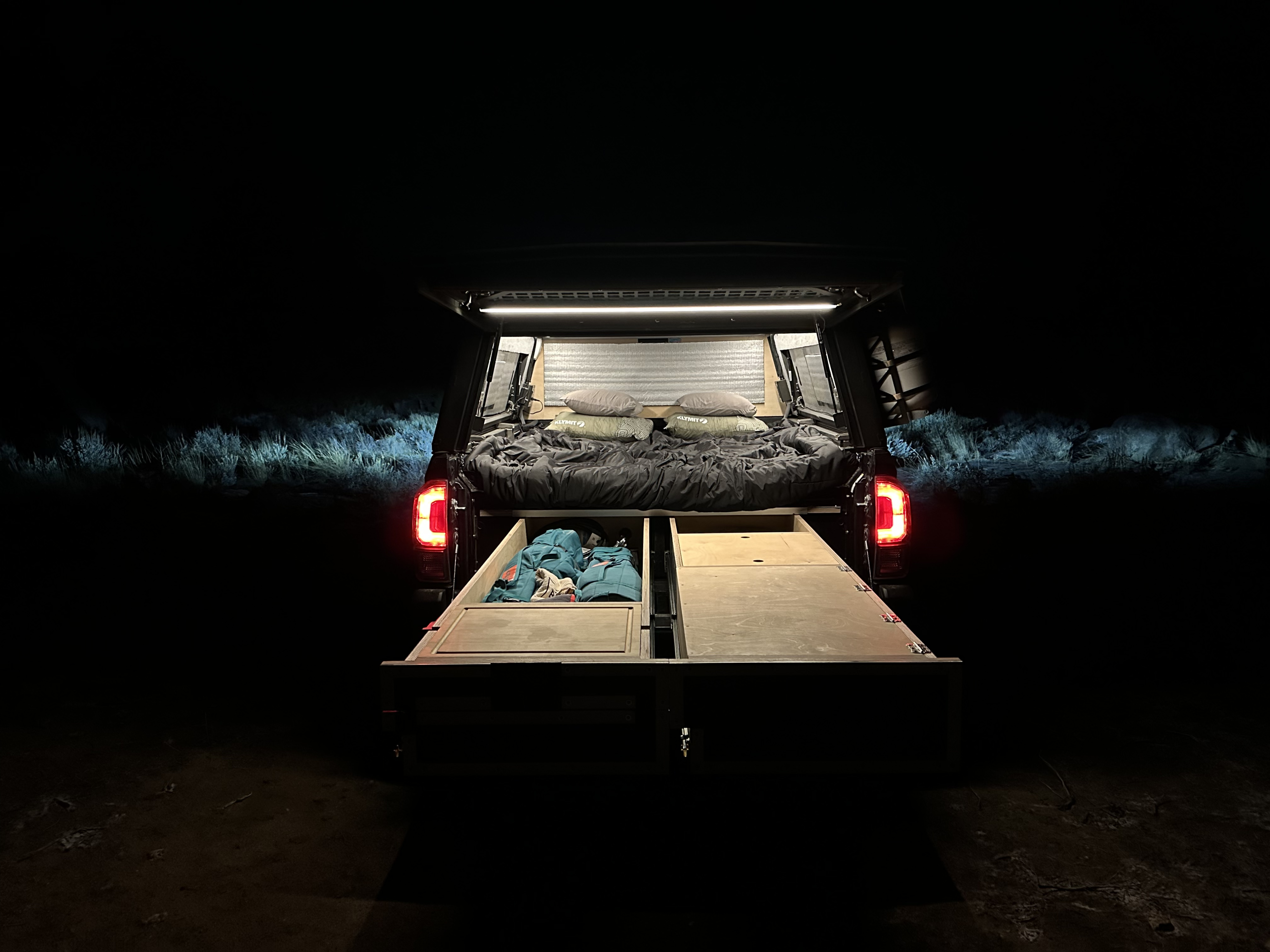 A cozy truck bed camping setup is lit at night, revealing a bed & storage drawers. Ideal for adventure, like an RV rental on wheels.. Alu-Cab Toyota Tacoma 2023