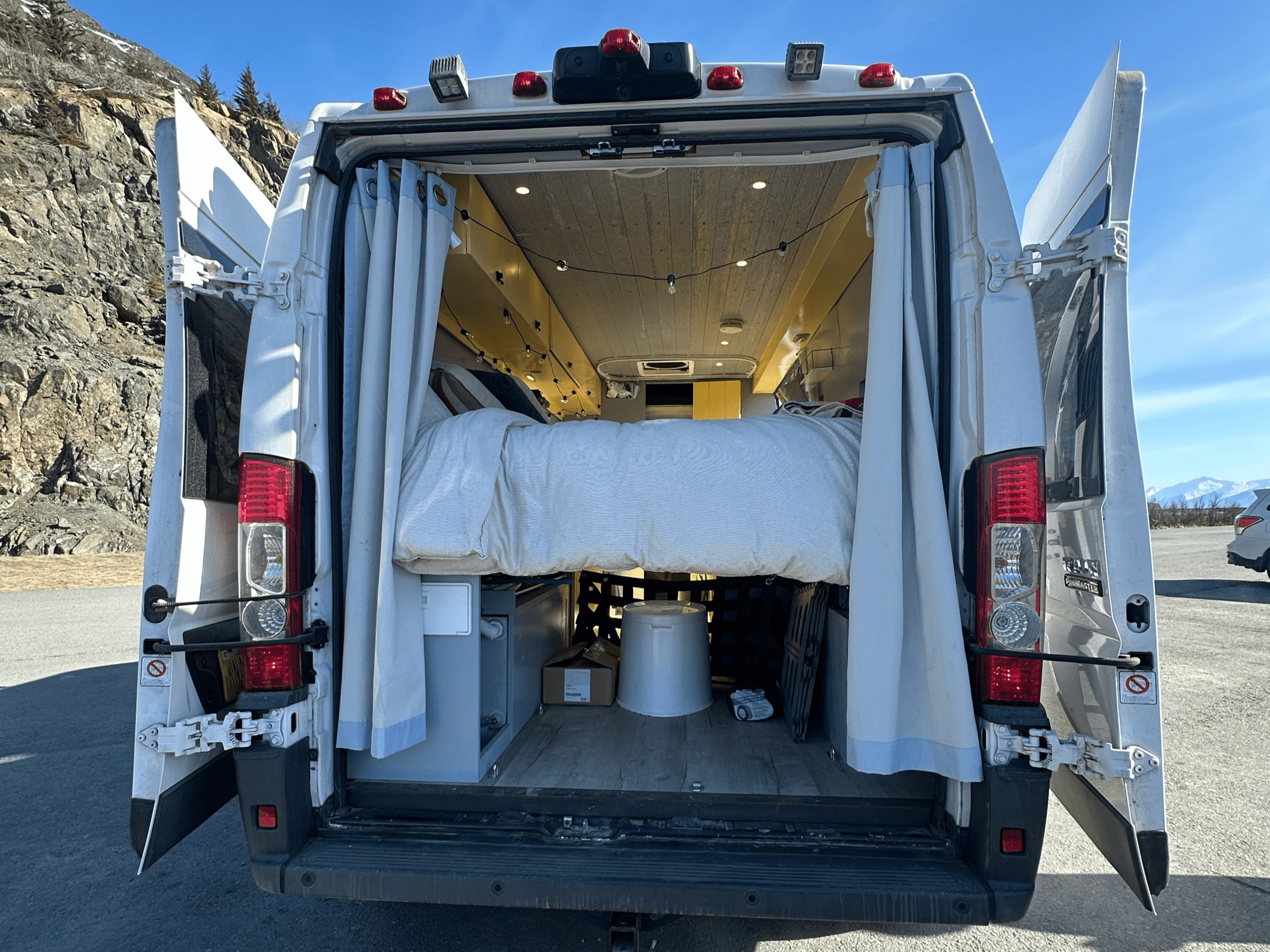 Open rear doors of an RV rental camper van reveal a cozy interior with a raised bed, lights, and under-bed storage.. Ram Promaster 2018