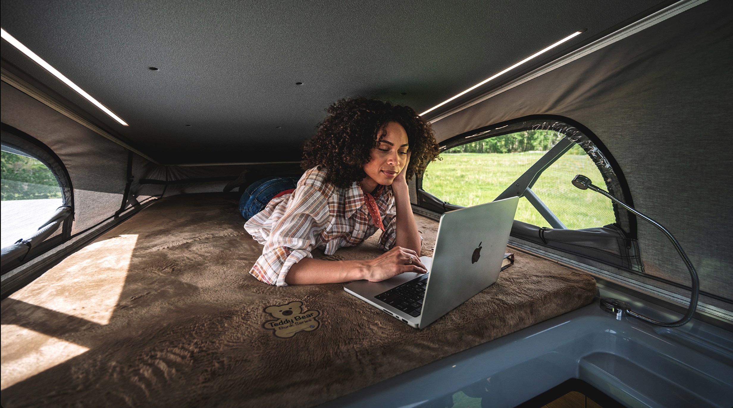A woman works on her laptop on a plush Teddy Bear bunk inside a pop-top rv rental, offering comfort with a view of nature.. Airstream Rangeline 2024