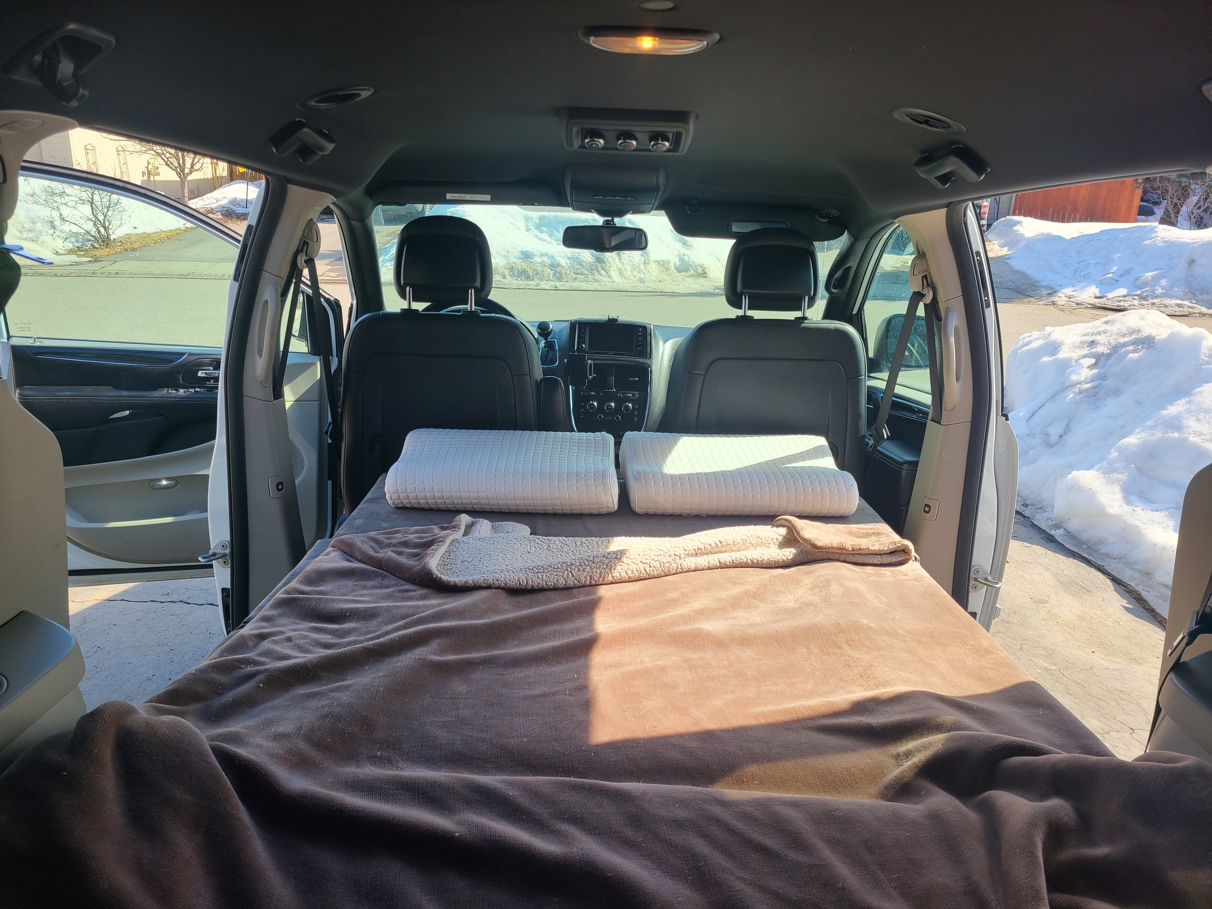 Inside a minivan, a cozy bed with pillows is set up for camping. An RV rental alternative ready for snowy adventures.. Dodge Mini C 2017