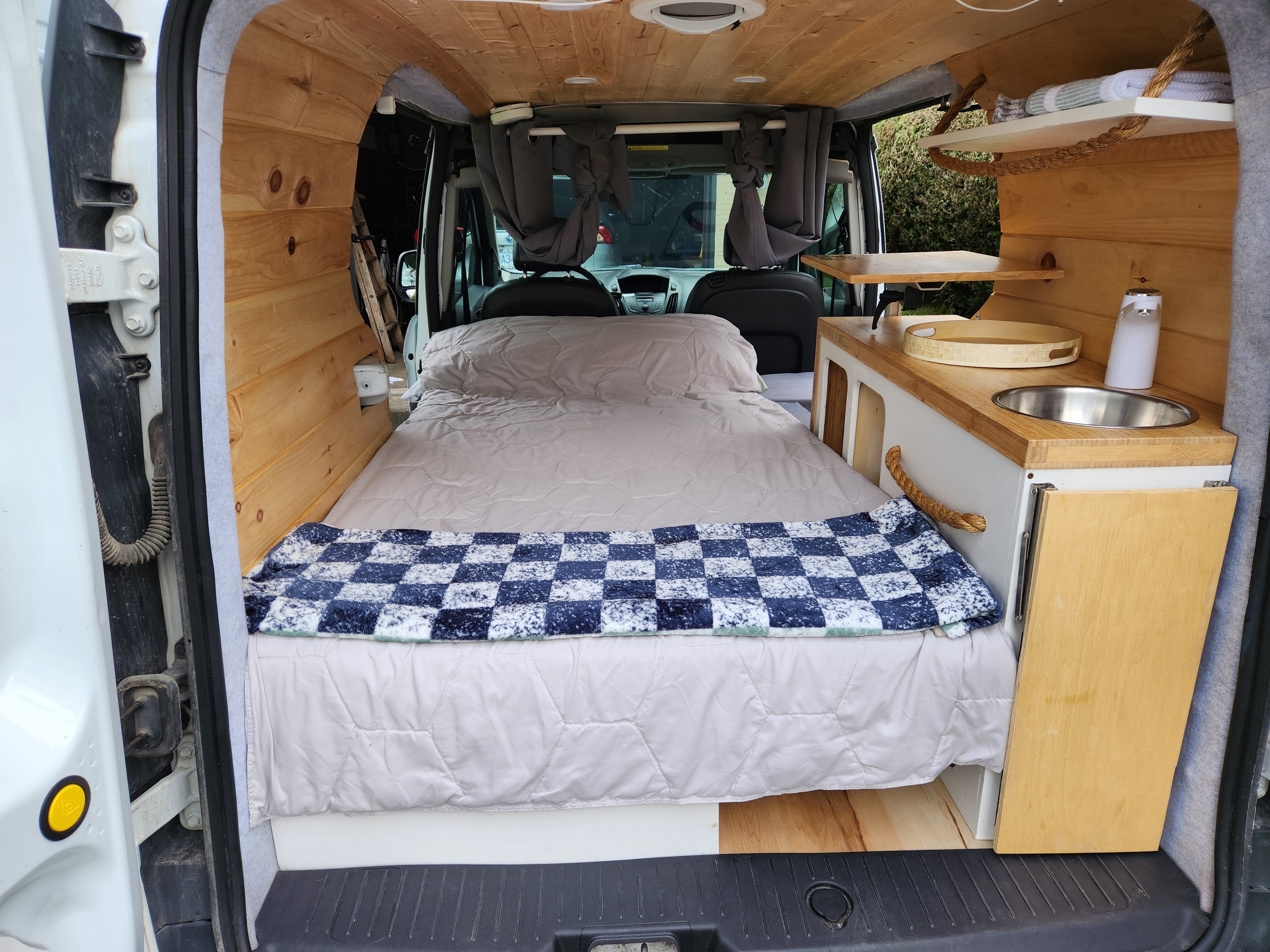 Cozy camper van interior from rear: bed, wood-paneled walls/ceiling, compact kitchen. Perfect for RV rental adventures.. Ford Transit 2015