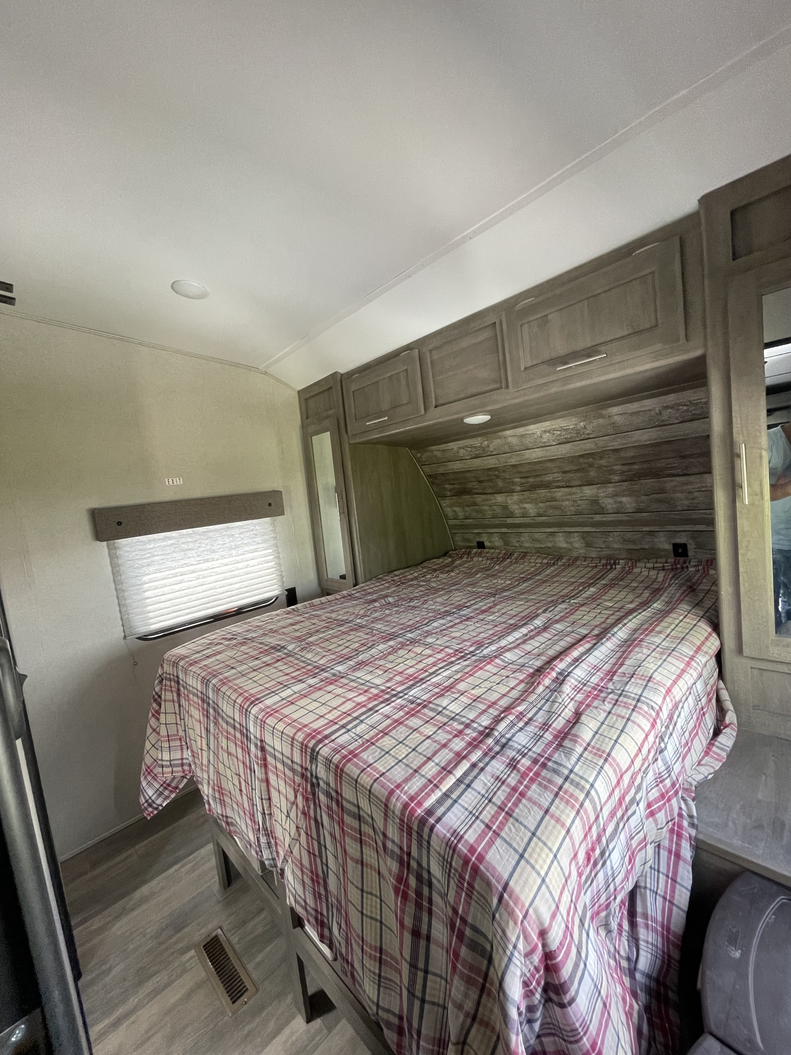 Cozy RV rental bedroom featuring a plaid bedspread, overhead storage cabinets, and a window with blinds. Ideal for comfortable travel.. Forest River XLR Boost 27QB 2021