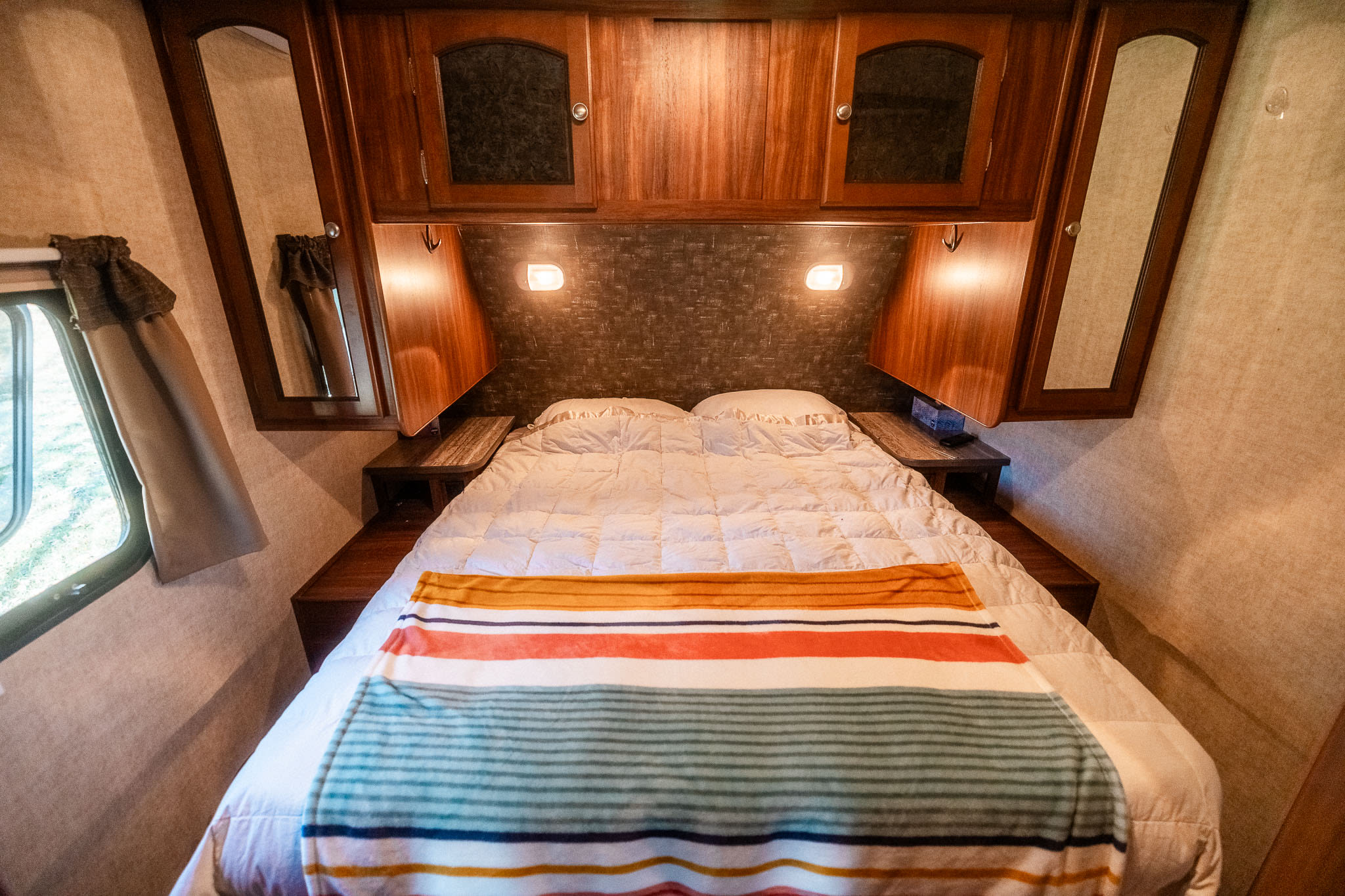 Experience comfort in this RV rental bedroom: bed with striped blanket, overhead storage, and warm lighting.. Heartland RVs North Trail 2015