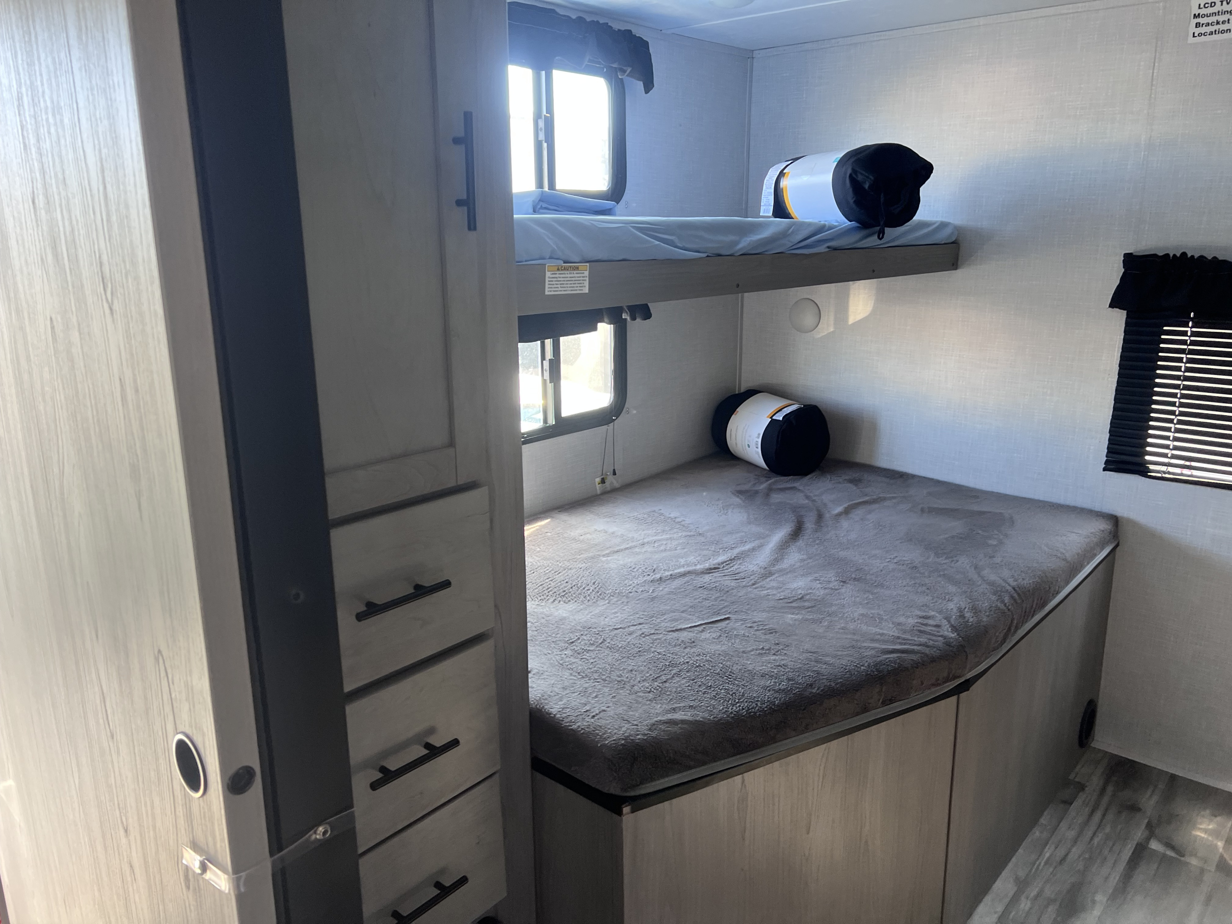 Interior view of an RV rental showing light-colored bunk beds, one with a window, and a slender dresser on the left.. Coleman 3215BH 2022