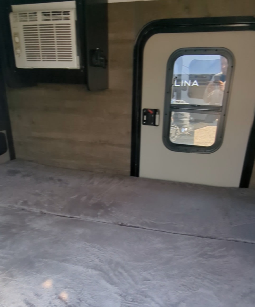 RV rental interior featuring a comfortable gray bunk, wall-mounted AC unit, and an entry door with a window.. Braxton Creek Bushwhacker 2023