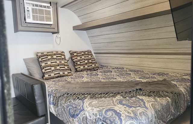 Comfortable RV rental bedroom featuring a bed with paisley bedding, accent pillows, an AC unit, and wood-paneled walls.. Dutchmen Coleman 2023