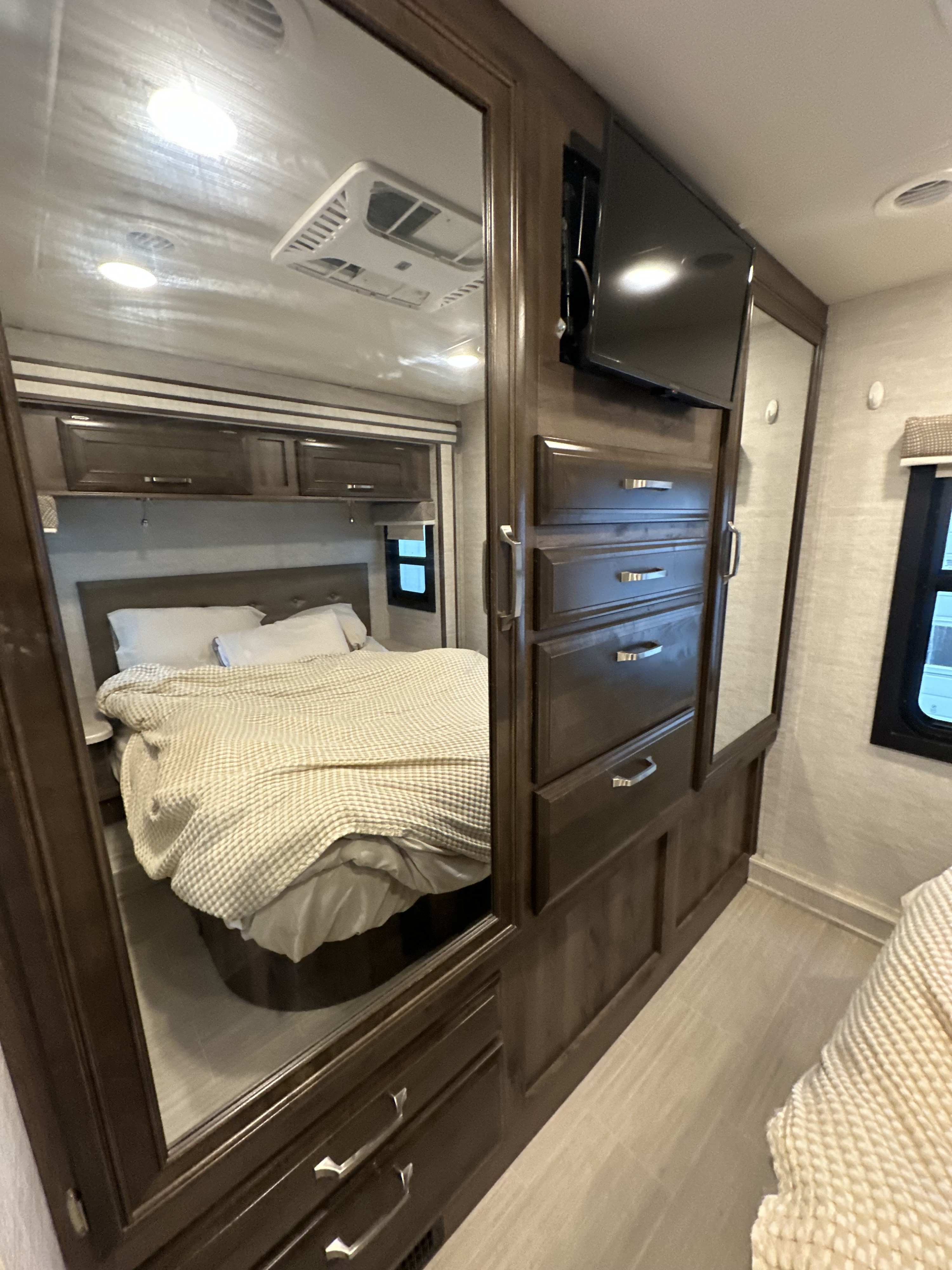 This RV rental bedroom features a comfortable bed, mirrored wardrobe, dresser with TV, and overhead storage cabinets.. Entegra Esteem 29V 2021