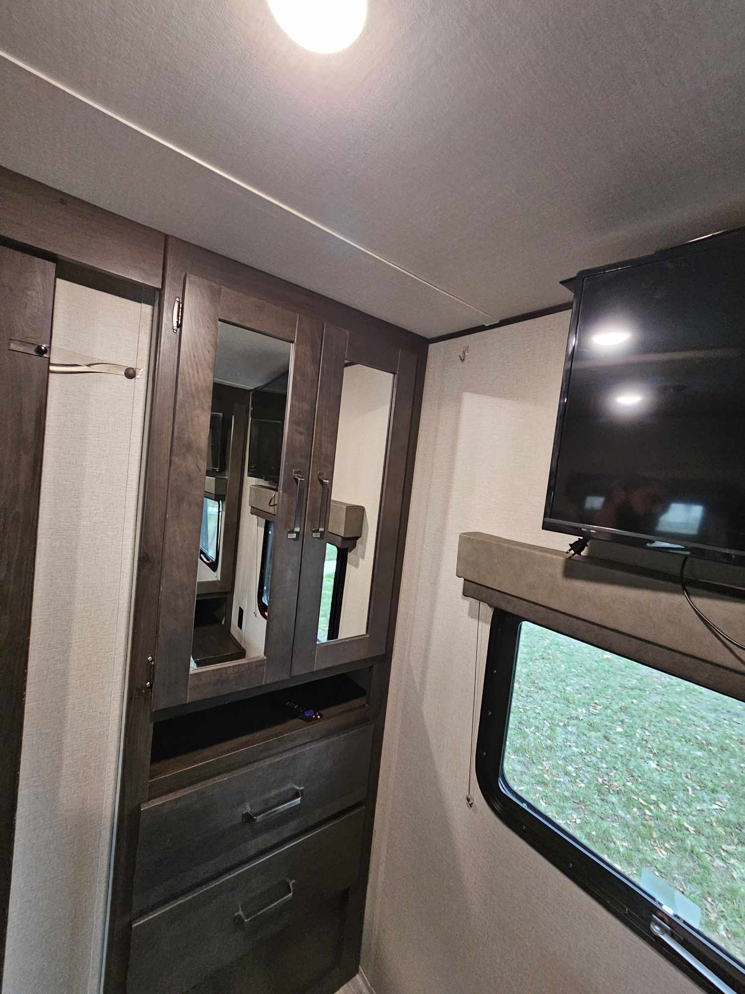 Inside an RV rental: built-in wardrobe with mirrors, wall-mounted TV, and window view of grassy outdoors.. Grand Design Transcend Xplor 2022